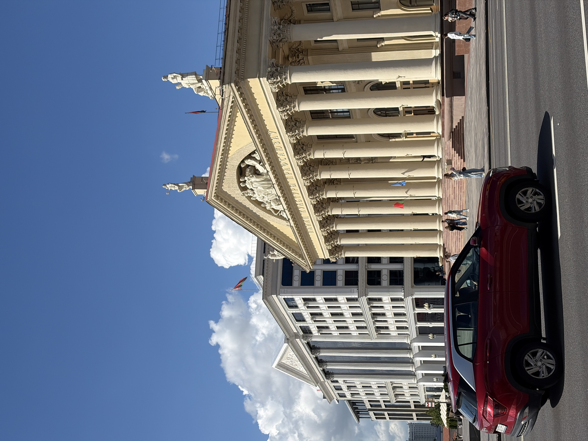 A low-angle, eye-level medium shot captures a red car driving on a paved road in front of two prominent neoclassical buildings under a bright blue sky with scattered white clouds. The building on the right is a pale yellow structure with a grand facade featuring a row of large Corinthian columns and a pediment adorned with sculptures. Above the pediment, two white statues stand against the sky. The building on the left is a modern white structure with multiple stories, large windows, and columns that echo the neoclassical style of the adjacent building. A flag flies from the rooftop of the building on the left. Several pedestrians are visible on the sidewalk between the road and the buildings, some walking and others standing. The red car, a compact SUV, is positioned in the foreground, moving from left to right. The overall scene depicts a sunny day in an urban setting, likely a prominent public square or avenue in Minsk, Belarus, given the architectural style and the flag.