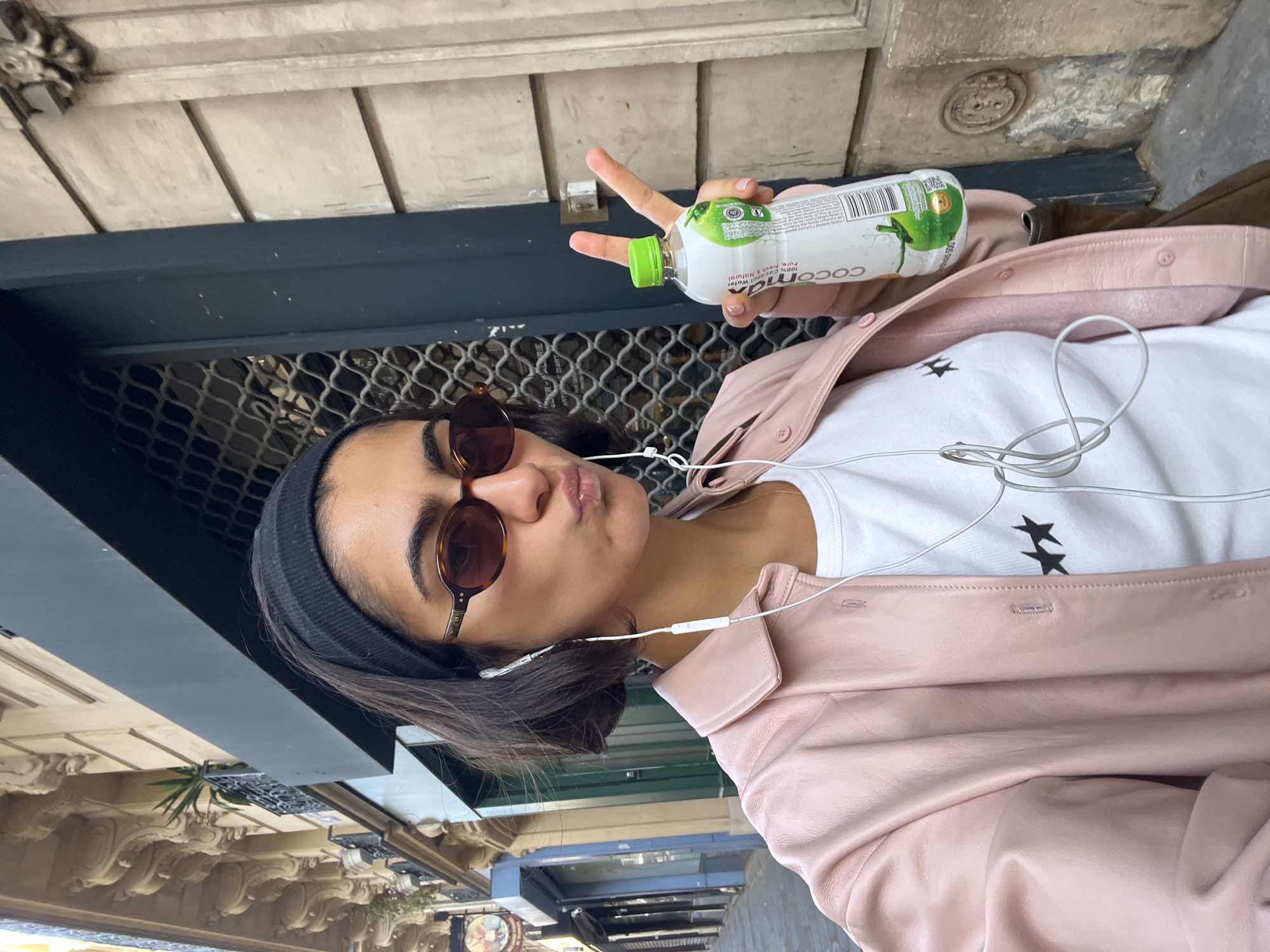 A young woman wearing a black headband, sunglasses, a white t-shirt with two black stars, and a pink leather jacket poses for a selfie outdoors, holding a bottle of coconut water and making a peace sign. She is wearing earbuds connected to a device that is not visible. The background shows Parisian architecture, including a metal grate and the facade of a building with ornate carvings. A store awning is visible in the distance. The scene appears to be a street in Paris, France.