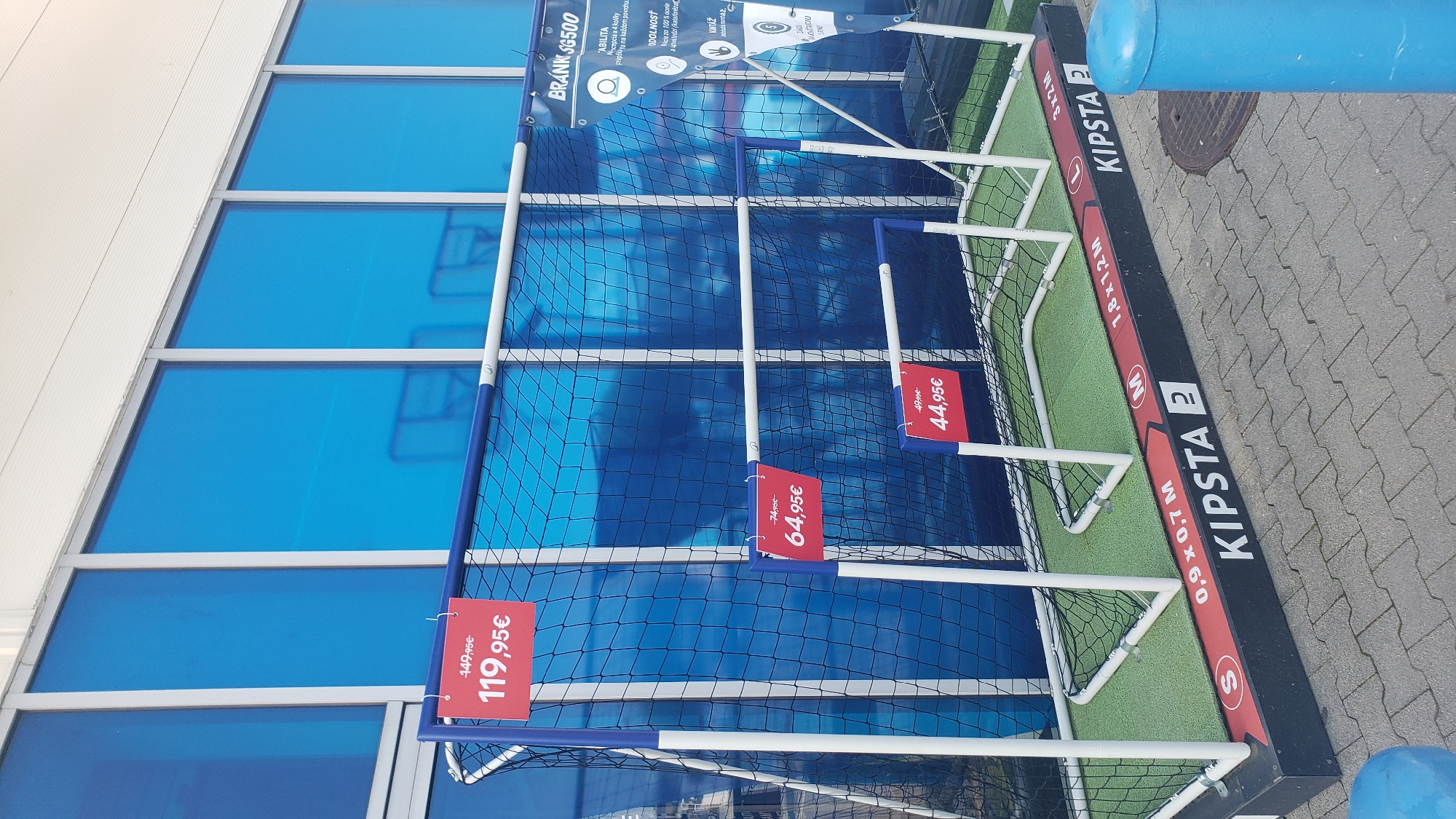 The image shows a display of soccer goals outside a store in Prešov, Slovakia. The goals are made of white and blue plastic tubing with black netting. There are three soccer goals of different sizes. Two are small and appear to be for children. The largest goal is in the background. Red price tags are attached to the goals. One price tag shows "149,95€" and "119,95€". Another price tag shows "74,95€" and "64,95€". A third price tag shows "49,95€" and "44,95€". The goals are placed on a green artificial turf surface. The background of the image is a blue glass building with white window frames. A blue cylindrical object is visible on the right side of the image. The ground in front of the display is paved with gray interlocking stones. The overall scene is an outdoor retail display.