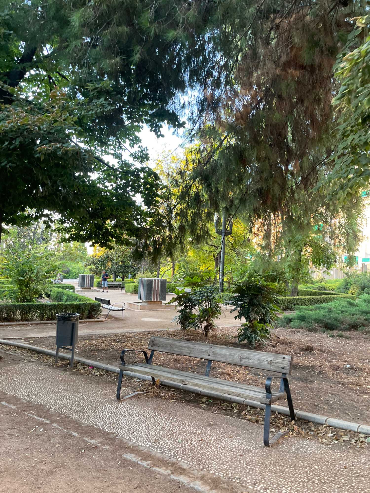 The image depicts an outdoor park scene, likely in Granada, Spain, given the mention of the city in the prompt. In the foreground, a weathered wooden bench with dark metal legs sits on a path paved with small, light-colored pebbles. To the left of the bench, a dark grey trash receptacle stands near the edge of the pebble path. The path itself is bordered by dirt and fallen leaves.
Behind the bench, the park opens up with manicured green hedges and shrubs. In the mid-ground, several large, dark rectangular structures with lighter colored marble accents are visible, possibly fountains or decorative elements. A lone figure, a person wearing a dark blue shirt and shorts, stands near one of these structures, facing away from the camera.
The background is filled with trees, some with dense green foliage and others with more wispy, pine-like needles, suggesting a mix of tree species. Sunlight filters through the leaves, creating dappled light and casting a warm glow, particularly in the upper right portion of the image. The overall atmosphere is serene and suggests a public green space within the city.