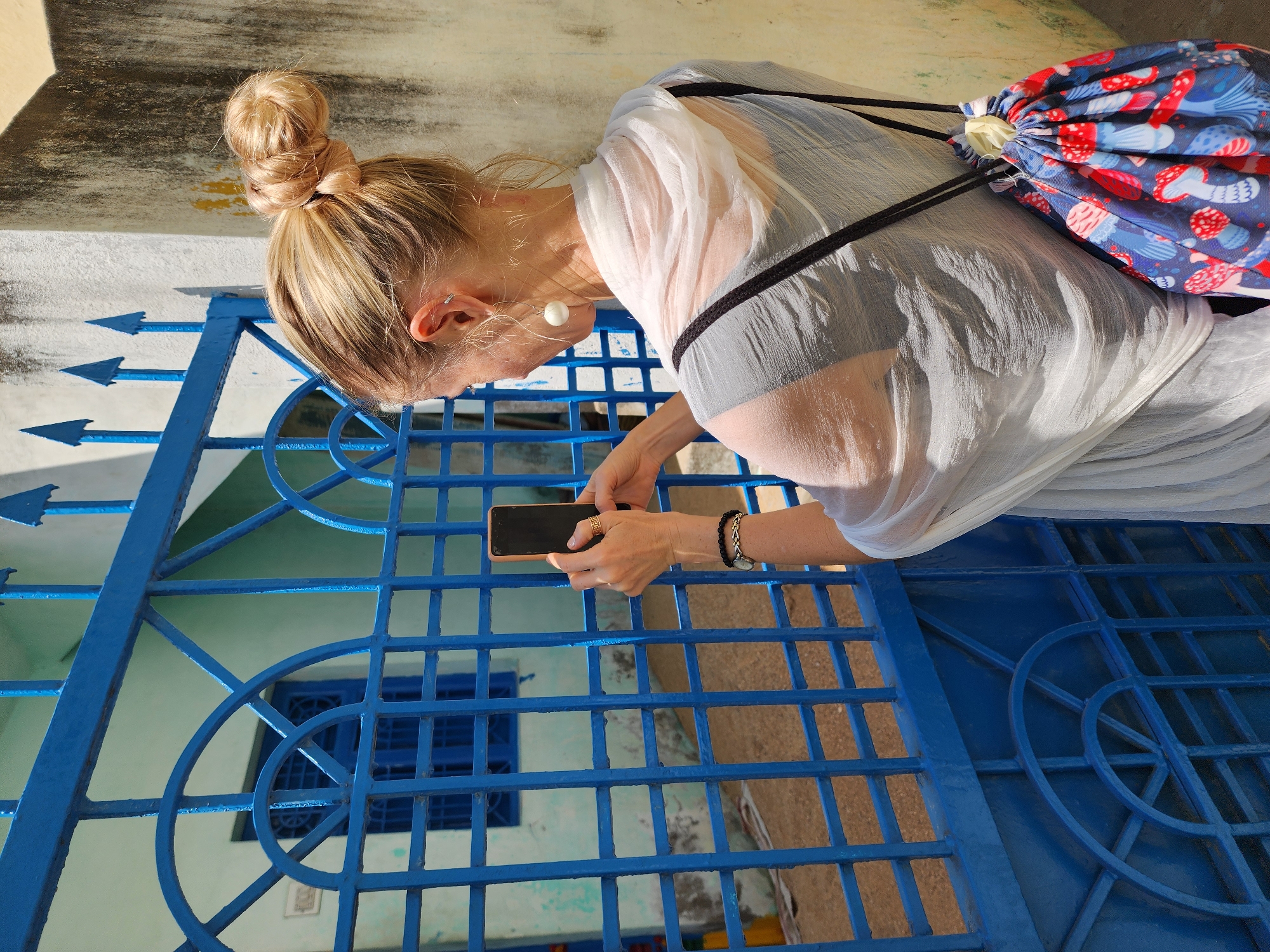 An individual, observed from the rear, with blonde hair styled in a high bun, stands facing a bright blue metal gate in Pedda Kammavaripalli, India. The person wears a translucent white shawl over a dark garment, white spherical drop earrings, and a blue drawstring backpack with a red and blue mushroom pattern. Both hands hold a dark-colored smartphone with a light orange or pink case, positioned to capture an image through the gate. The blue metal gate features a grid pattern, decorative arched elements, and pointed finials along its top edge. Beyond the gate, a light blue or pale green building is visible, featuring a window with blue grates and a dark blue door, also with grates. The ground visible through the gate appears to be dry and light brown. To the right, a weathered, light-colored wall with dark streaks and yellow discoloration runs vertically, providing a backdrop to the scene.