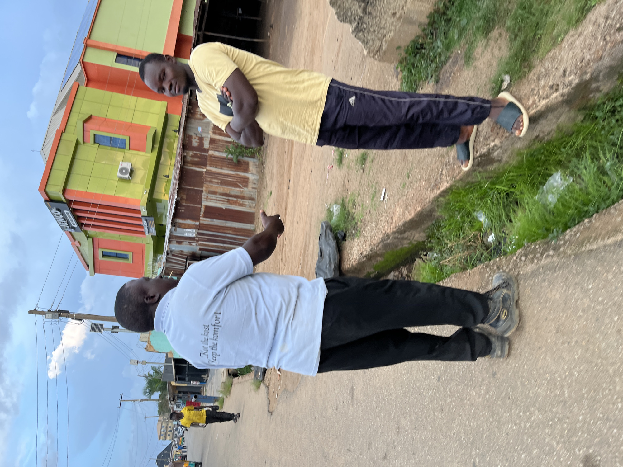 The image, correctly oriented, displays two dark-skinned men in the foreground engaged in an interaction on an outdoor street or pathway in Jos, Nigeria, with a vibrant building and urban elements in the background.
The man on the left wears a light yellow t-shirt and dark blue or black track pants with white side stripes, complemented by light-colored sandals. He stands with his arms crossed, looking towards the other man. His short hair is neatly cut.
The man on the right is viewed from behind, wearing a white t-shirt with the phrase "Not the best / Keep the komfort" printed in black text. He has dark pants and dark sneakers and appears to be mid-gesture with his right hand, facing the man in yellow.
In the background, a striking multi-story building features bright green and orange/red cladding, with blue-framed windows and an air conditioner unit on its exterior. Solar panels are visible on its roof. A sign on the building's facade partially reads "HENCO". Adjacent to this building is a rusty corrugated metal fence or structure.
The ground beneath the men consists of a paved surface, which transitions to a dirt or gravel area and then a strip of grass with a drain on the far right, where some litter, including a plastic bag, is visible. Utility poles and overhead wires crisscross against a bright, partly cloudy daytime sky. Another person in a yellow shirt and dark pants is seen further down the street in the distance. The scene suggests a busy, urban environment during daylight hours.