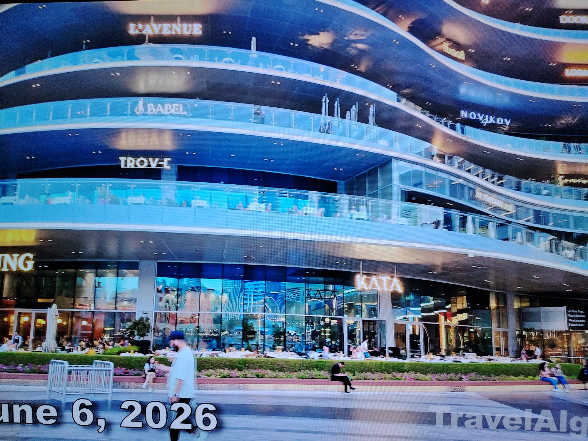 This is a medium-distance, eye-level, slightly tilted, wide-angle shot of a modern, multi-level building with curved facades and glass balconies. The building has several levels with illuminated signs displaying various brand names such as "L'AVENUE", "BABEL", "TROV-I", "NOVIKOV", and "KATA". The exterior is predominantly blue and reflective, with glimpses of interior lights and architectural details.
In the foreground, a blurred street scene is visible. The date "June 6, 2026" is superimposed in large, white, sans-serif font. A watermark reading "TravelAlg" is also present in the lower right corner.
Several individuals are present on different levels of the building and on the street. On the ground level, patrons are seated at outdoor cafes. On the street level, people are walking and sitting. One person in the foreground is walking away from the camera, wearing a blue cap and white t-shirt. Another individual is sitting on a low wall, looking at their phone.
The environment appears to be urban, with a focus on the architecture and signage of the commercial building. The lighting suggests it is daytime, but the reflections and internal lighting of the building create a mixed illumination. The location is not specified as Huntingdonshire, United Kingdom, in the image.