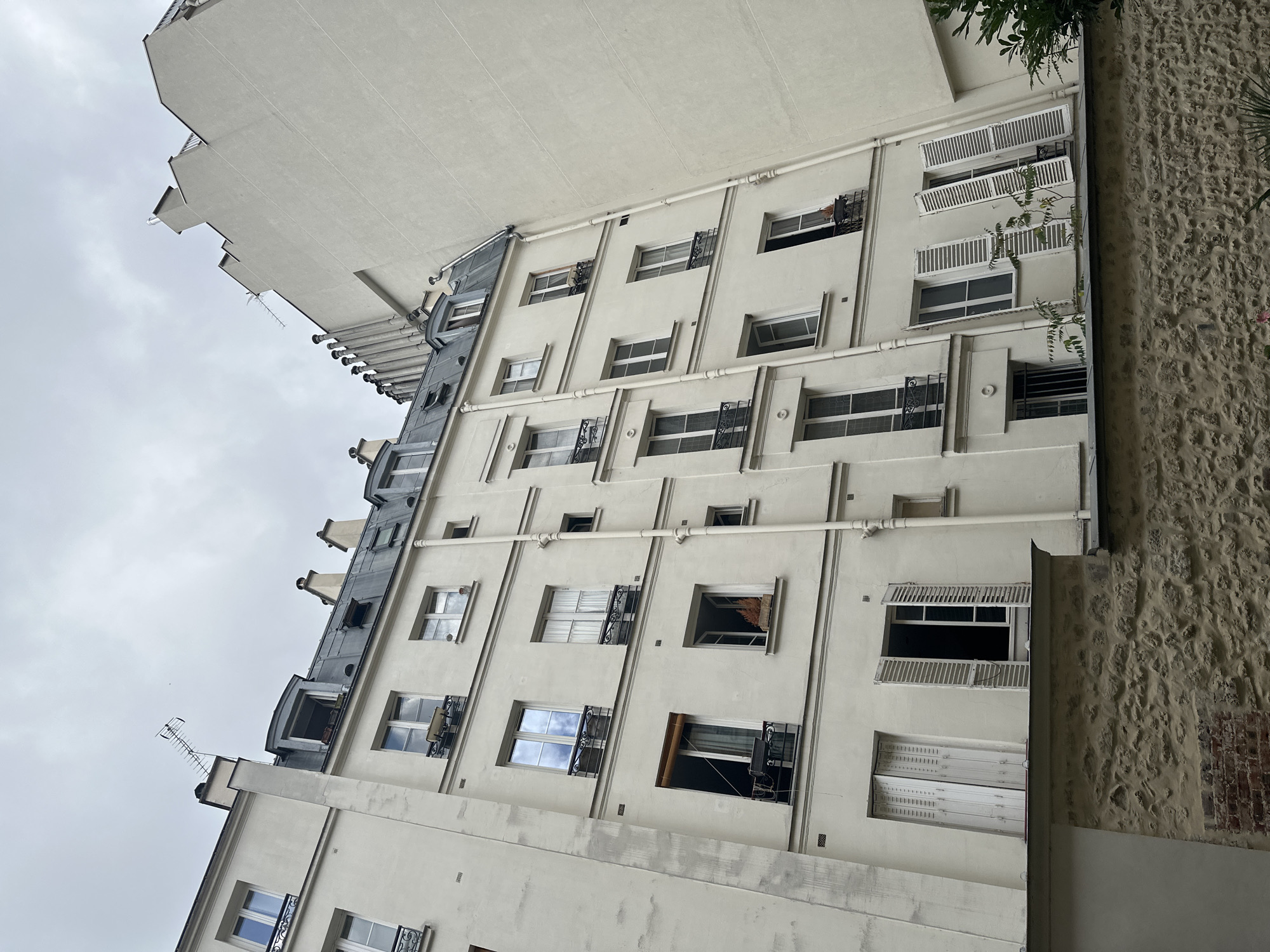 The image depicts a section of a multi-story building in Paris, France, likely in a residential area given the architecture. The building features a pale, off-white facade with numerous windows, many of which are adorned with white shutters. Some windows are open, revealing glimpses of the interior, while others are closed. The top of the building showcases a grey, mansard-style roof with dormer windows and chimneys. The sky above is overcast and grey. In the foreground, a stone wall runs horizontally across the lower part of the image, partially obscuring the base of the building and suggesting a courtyard or lower garden area. Some greenery is visible in the bottom right corner, suggesting the presence of trees or plants. The overall impression is one of a typical Parisian Haussmannian building, common in many arrondissements of the city. No people are clearly visible in the image.