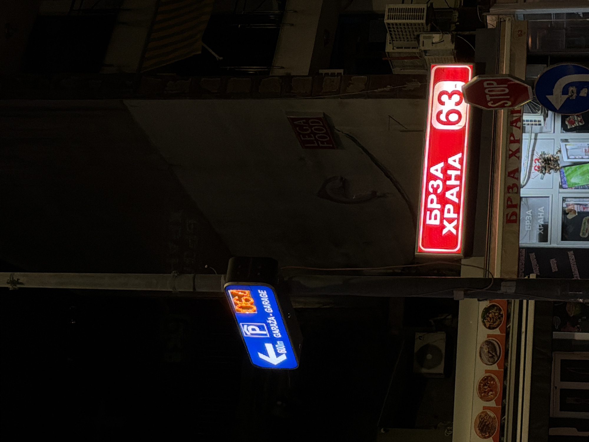 The image captures a street scene at night, likely in Trebesin, Montenegro, based on the mix of Cyrillic and Latin script on the signage. The scene is dominated by several illuminated signs:
1. A red vertical sign reading "63 БРЗА ХРАНА" (Fast Food).
2. A sign for a fast-food restaurant, with the Cyrillic "БРЗА ХРАНА" (Fast Food).
3. A "STOP" sign.
4. A blue sign indicating a garage ("GARAŽA-GARAGE") 600 meters away with the designation "067 P" indicating possibly a parking location.
5. "LEGA FOOD" is visible on a sign.
The surrounding environment appears to be a business district or commercial area, judging from the storefront and signage. There is a street lamp or other light source casting light on the signs. The overall atmosphere is typical of a nighttime urban scene.