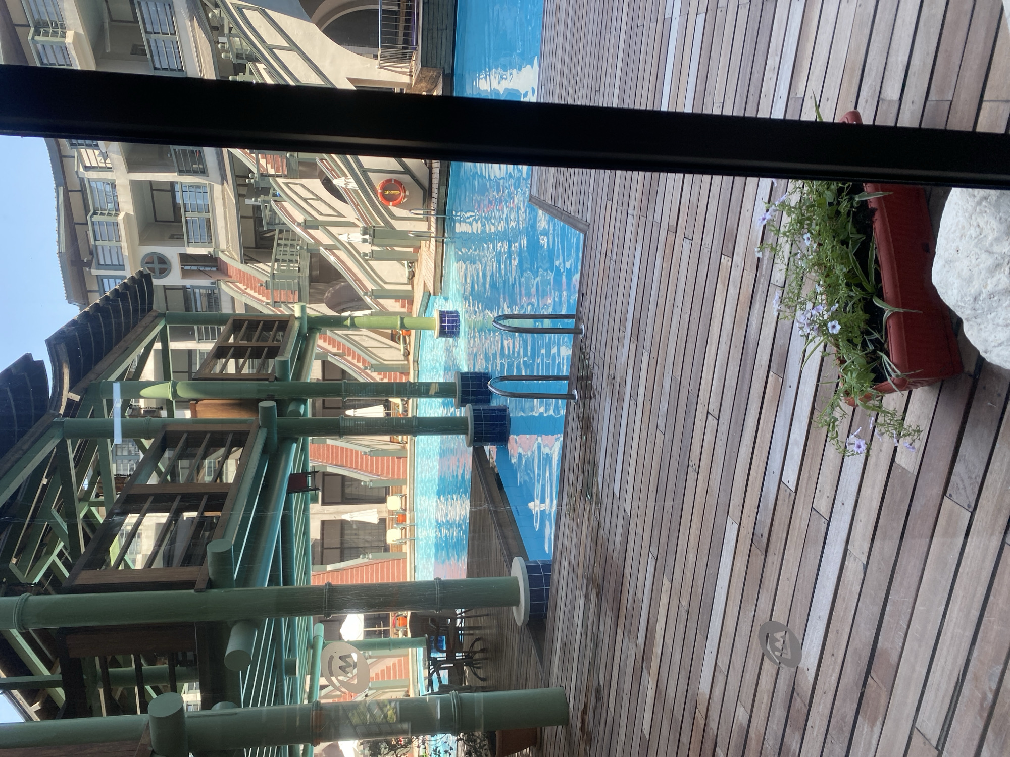 The image shows a swimming pool area at a resort. In the foreground, a wooden deck leads to the pool. A red planter with purple flowers sits on the deck. The pool is a bright blue and has a set of silver pool stairs. In the background, a multi-story building with balconies and staircases overlooks the pool. The architecture suggests a tropical or Mediterranean style. The scene is set in Aksu, Turkey, likely at a hotel or resort complex. There are no people visible in the image. The sky is clear and blue.