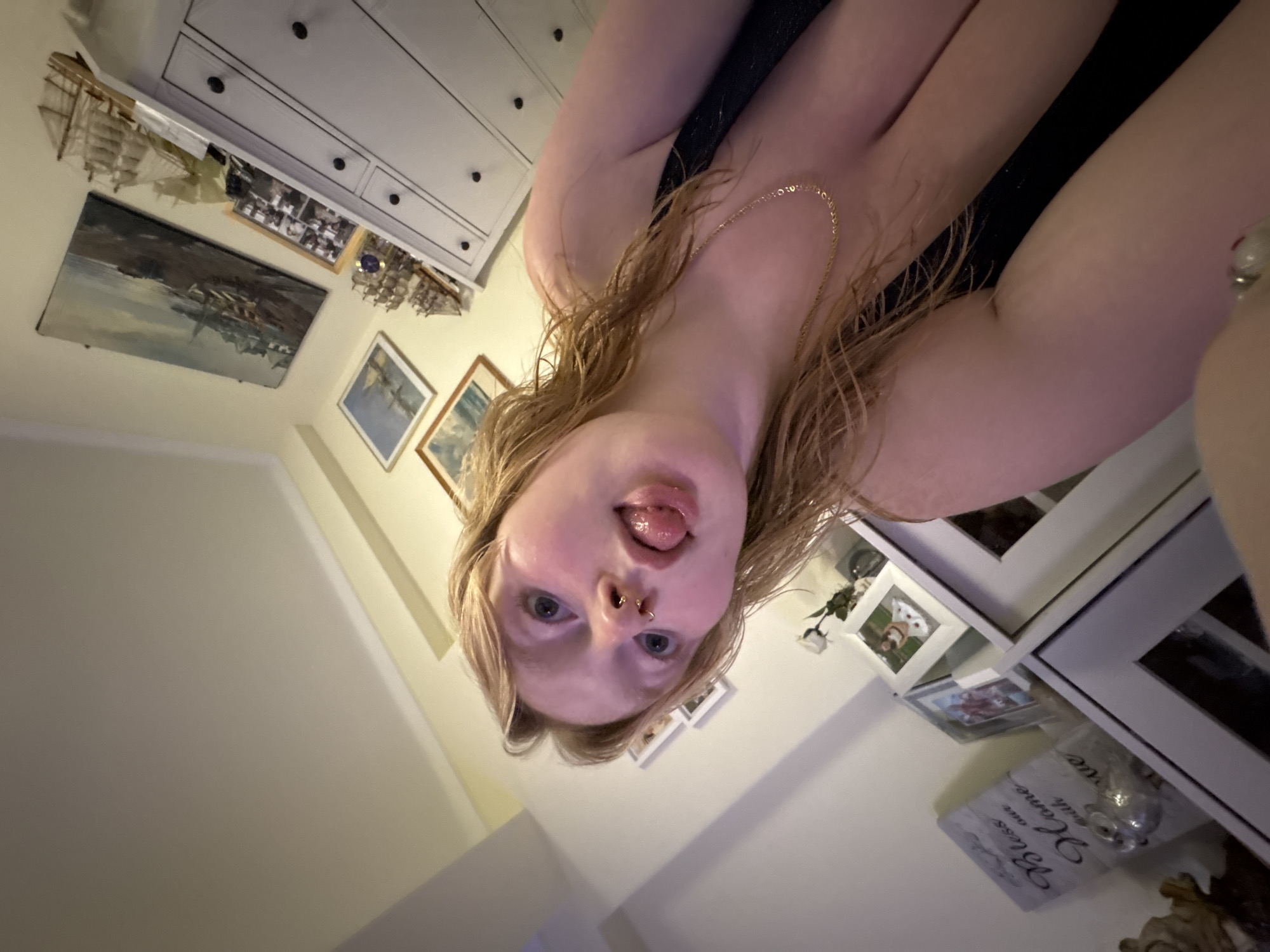 A low-angle indoor shot features a woman with fair skin and wet, blonde hair. She has blue eyes and a pierced nose with two rings. Her tongue is sticking out. She is wearing a thin, gold necklace and a dark-colored garment.
The background reveals a portion of a white dresser with black knobs. Above the dresser, there are several framed artworks, including paintings of seascapes and model ships. There's also a framed print that says "Bless our Home with Love." The walls are painted in a pale, warm tone.