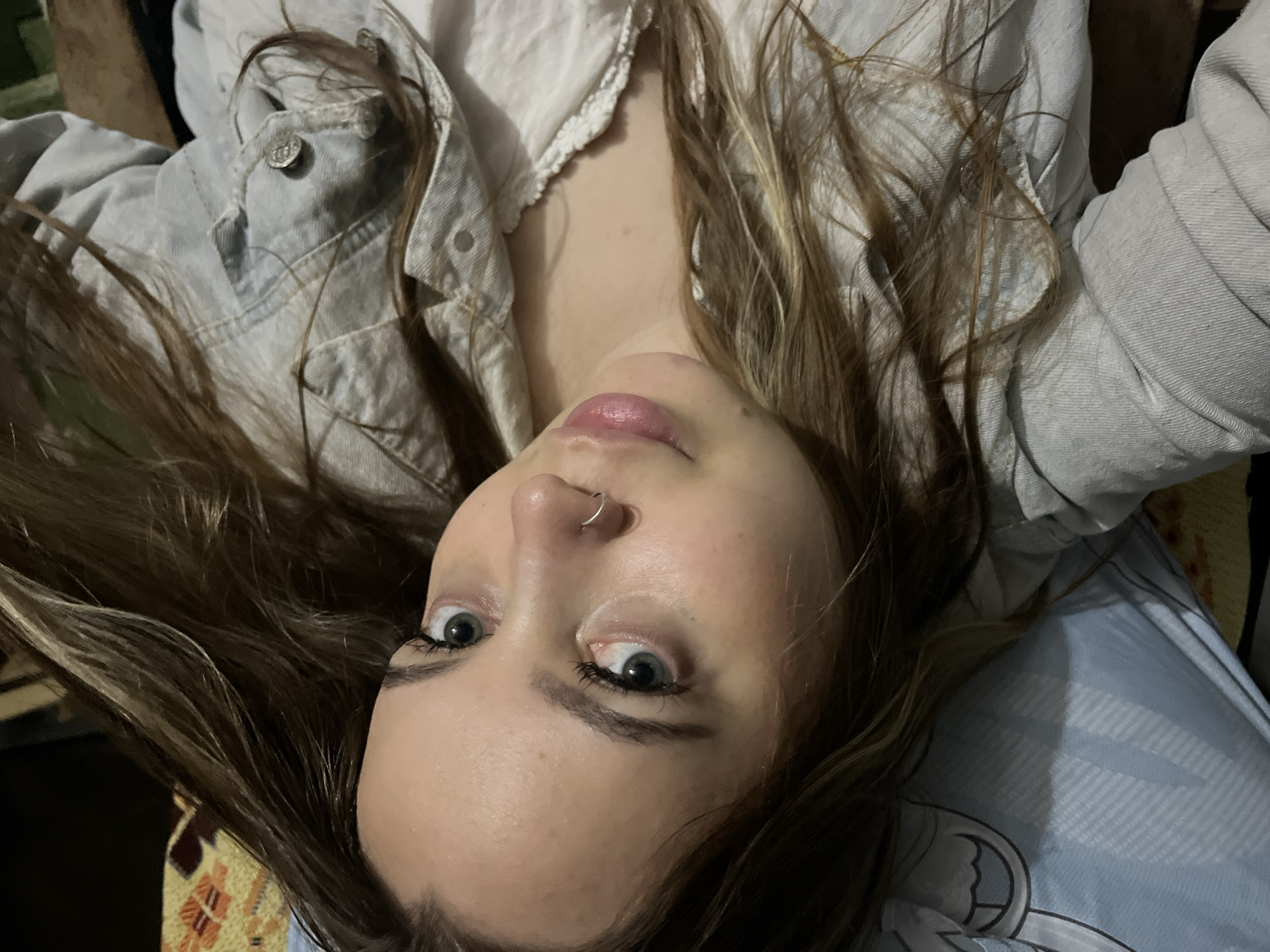 A young woman with brown hair and light blue eyes is lying down. She has a silver nose ring. She is wearing a light blue denim jacket over a white shirt with a frilly neckline. The background includes patterned fabric and dark areas. The image appears to be a selfie taken from above. There is no information in the image to indicate a location in Novopokrovskaya, Russia.