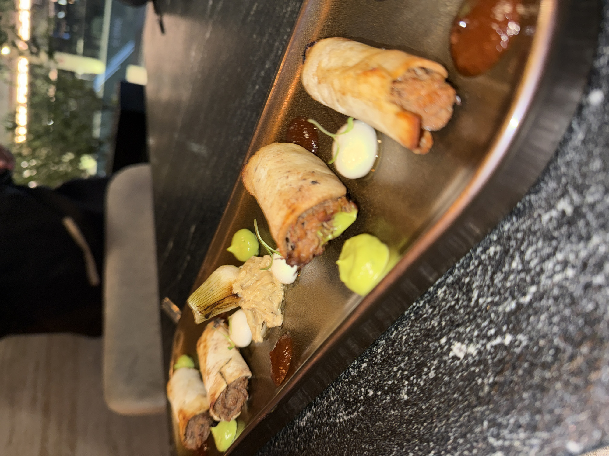 The image displays a close-up, angled view of an appetizer or small plates serving on a dark, rectangular tray, likely in a restaurant or bar setting during the evening. The tray holds approximately five small, cylindrical food items, resembling rolls or taquitos, filled with a savory, brownish mixture.
These food items are accompanied by several dollops of various sauces and garnishes. There are bright green dollops, possibly an avocado or herb emulsion, as well as smaller white and reddish-brown dollops, likely creams or relishes. A piece of roasted or blanched vegetable, possibly a leek or spring onion base, and a clump of shredded or braised vegetable or fungus are also arranged on the tray.
The background is significantly blurred, with a person partially visible on the left side of the frame, wearing dark clothing. Beyond them, through a window, out-of-focus lights from an outdoor setting, such as a city view or decorative lighting, are discernible, indicating a nighttime environment. The table surface appears to be a dark, speckled material, possibly granite or marble. No text is visible in the image.