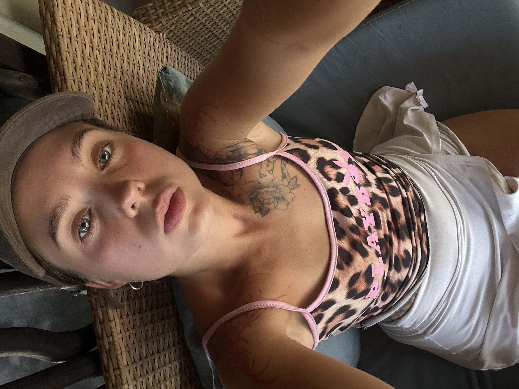 A woman with blonde hair and blue eyes is lying down and looking at the camera. She is wearing a baseball cap, a leopard-print tank top with pink trim and the word "PLAYBOY" in pink lettering, and a shiny, light-colored skirt. She has tattoos on her arms and shoulders. The background shows a woven chair and some dark wood structures. The lighting suggests it is daytime.