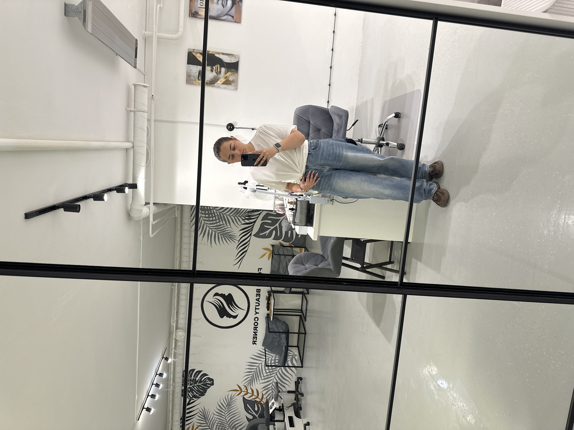A woman stands in a well-lit room, taking a mirror selfie with her phone. She is wearing a white t-shirt and light-wash denim jeans, with brown shoes. The room appears to be a beauty salon, evidenced by the decor, which includes wall art featuring abstract faces and botanical prints. Several styling chairs and treatment stations are visible in the reflection. The floor is a light gray speckled material, and the ceiling has track lighting and ventilation systems. The overall aesthetic is modern and clean. The location is identified as Shchyolkovo, Russia.