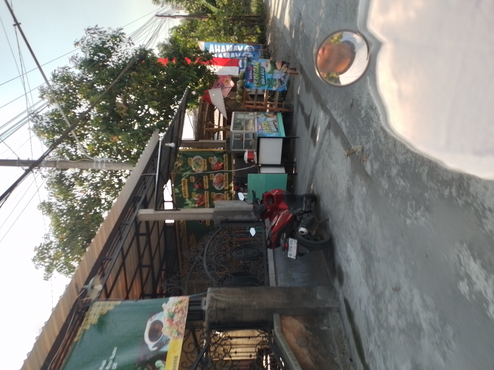In Wates, Indonesia, a red motorcycle is parked on a paved area in front of a food stall. The stall has a corrugated metal roof supported by metal beams, and a large black metal gate with decorative scrollwork is visible behind the motorcycle. Various signs are displayed at the stall, including one that advertises "ES DEGAN GULA ASLI" (Authentic Palm Sugar Coconut Ice). To the right of the stall, a wooden stand holds several coconuts. Red and white Indonesian flags are also displayed. Trees and power lines are visible in the background. A circular mirror on the ground reflects the sky and a person's face. The scene appears to be a street-side establishment on a clear day.