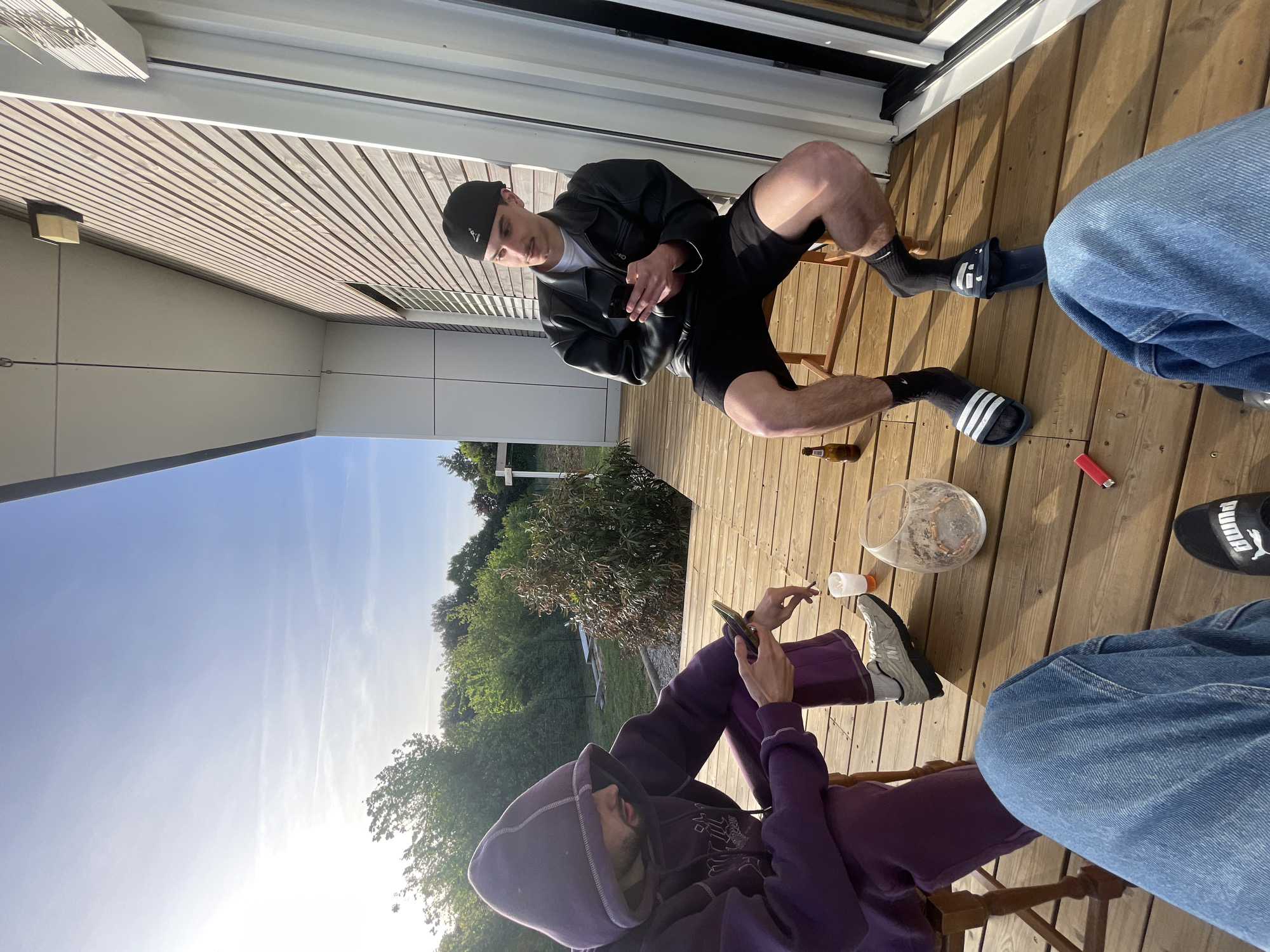 This image, taken in Thénezay, France, depicts a casual outdoor scene on a wooden deck, likely during the late afternoon or early evening given the bright, low sun.
Two men are seated on wooden chairs on the deck. The man on the right (when oriented correctly) is wearing a black leather jacket over a white t-shirt, black shorts, a black cap with a small white logo, black socks with a white Nike swoosh, and black Adidas slides featuring the iconic three stripes. He is holding and looking at a smartphone. The second man, positioned in the foreground, is wearing a purple hooded sweatshirt and matching sweatpants, along with grey New Balance sneakers. He is also looking at a smartphone. Partially visible in the lower right foreground are the legs of a third person, wearing blue jeans and a black Puma slide with the brand's logo.
Scattered on the wooden deck are several items, suggesting a relaxed social setting: a glass bowl containing ash and cigarette butts, a red lighter, a small white plastic cup, and a brown beer bottle.
The background reveals a sunny day with a clear blue sky, showing some faint contrails. Lush green trees and bushes are visible beyond the deck, suggesting a natural or garden environment. Part of a modern building with light-colored horizontal siding is also visible to the left. The overall mood appears relaxed and informal.