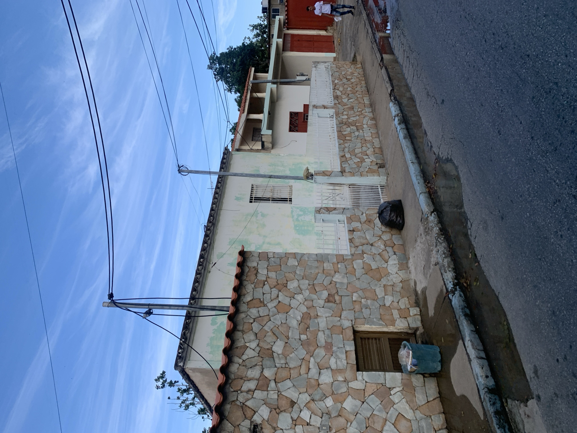This is a street-level, medium shot image captured in La Asunción, Venezuela. The scene depicts a residential street with several houses visible. In the foreground, a gray asphalt road shows patches of standing water, indicating recent rain or drainage issues. A sidewalk runs alongside the road, bordered by a curb.
On the left side of the image, a house features a lower section clad in multi-colored, rough-cut stone. Above this, the house wall is painted a pale green, with visible signs of wear and tear. A section of white metal fence runs along the front of the property. A teal trash bin filled with garbage is placed near the stone wall. To the right of this house, a white metal fence encloses the property, and a black trash bag lies on the sidewalk behind it.
Further to the right, another house is visible, painted in lighter colors. It has a second story with a balcony supported by pillars. Red doors are prominent on this property, one of which is large and opens to the street. A man in a white t-shirt and blue jeans is walking away from the camera on the sidewalk, holding a white object in his right hand.
Overhead, a network of electrical wires crisscrosses the sky, supported by utility poles. The sky is a pale blue with wispy white clouds. Trees are visible behind the houses, adding to the urban residential setting. The overall impression is of a typical street scene in a city with older architecture and some signs of aging infrastructure.