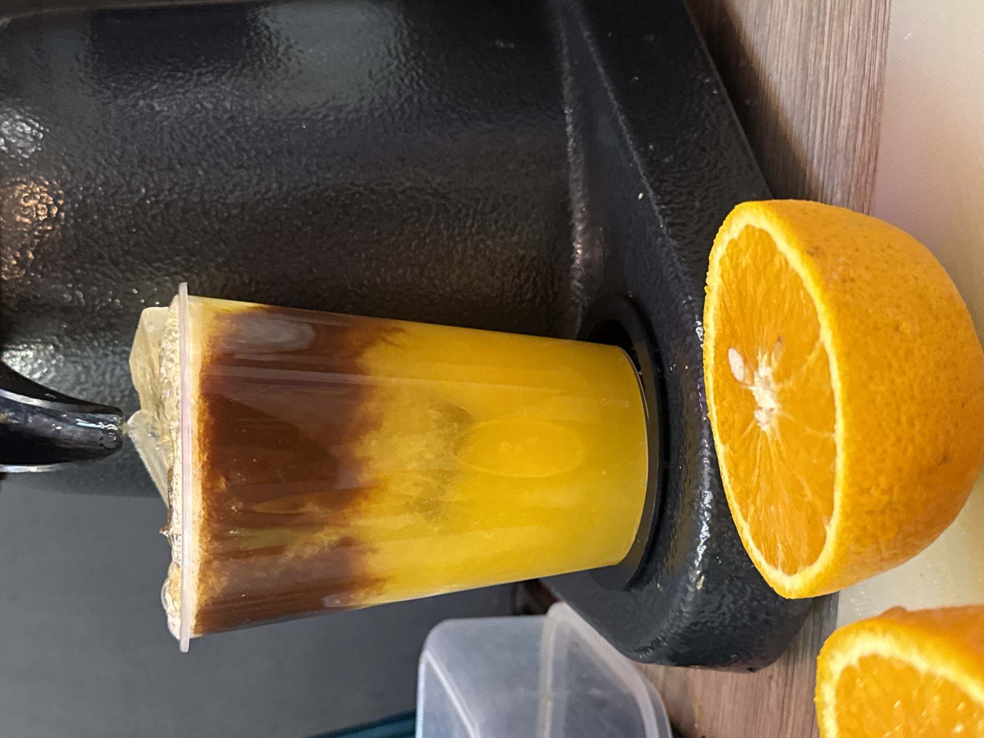 A layered drink, likely coffee and orange juice, sits in a clear plastic cup on a black base, with a sliced orange in the foreground. The drink is being dispensed from a black machine into the cup. The scene appears to be set on a wooden surface, with a gray background. No people are visible.