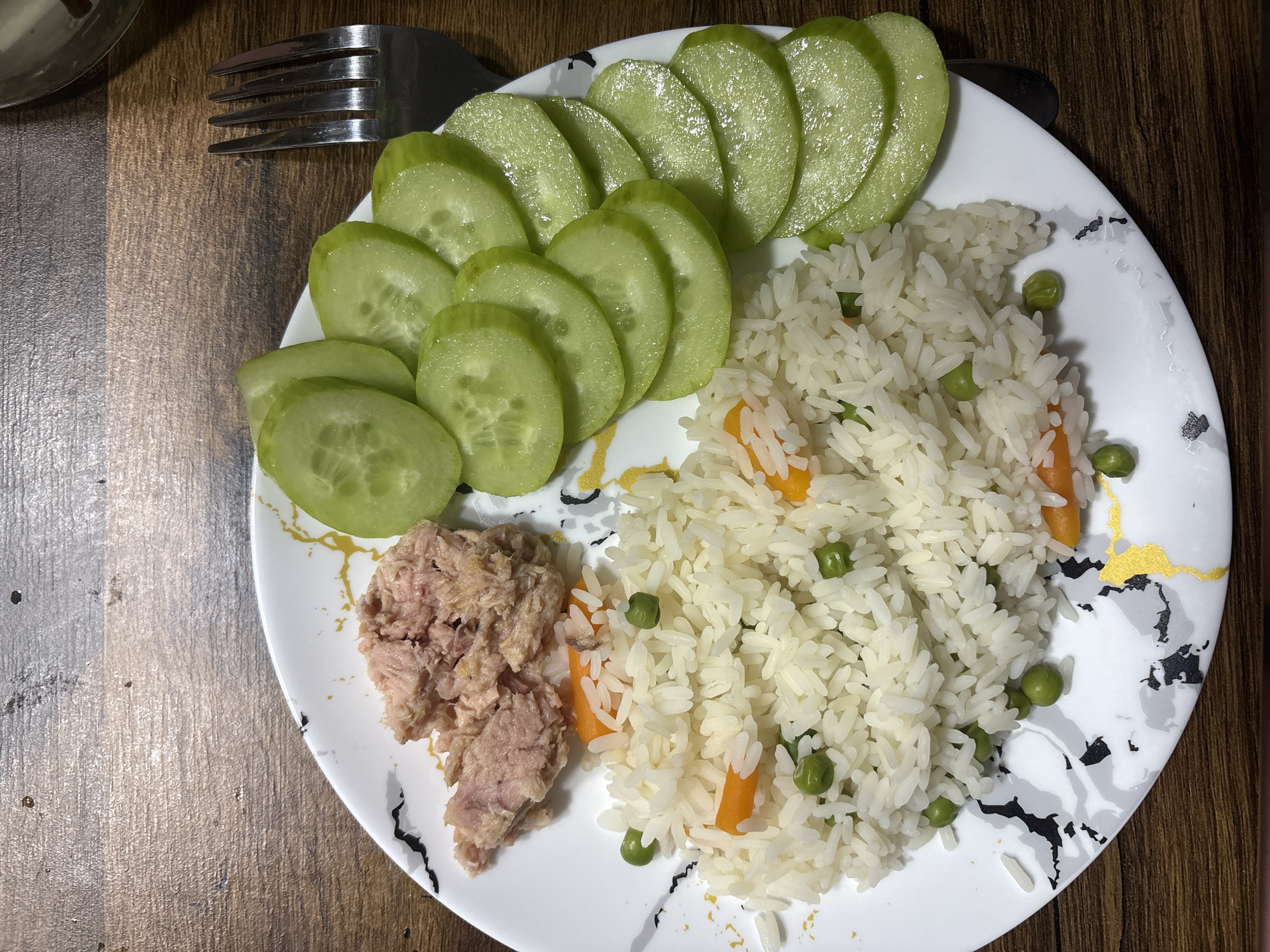 A plate of food is presented on a wooden table. The plate is white with a marble-like pattern of black and gold. On the plate, there is a pile of white rice mixed with green peas and chopped carrots. To the left of the rice is a serving of canned tuna. To the right of the rice are several slices of cucumber arranged in a fan shape. A silver fork rests on the right side of the plate. The background is a wooden surface, likely a table.