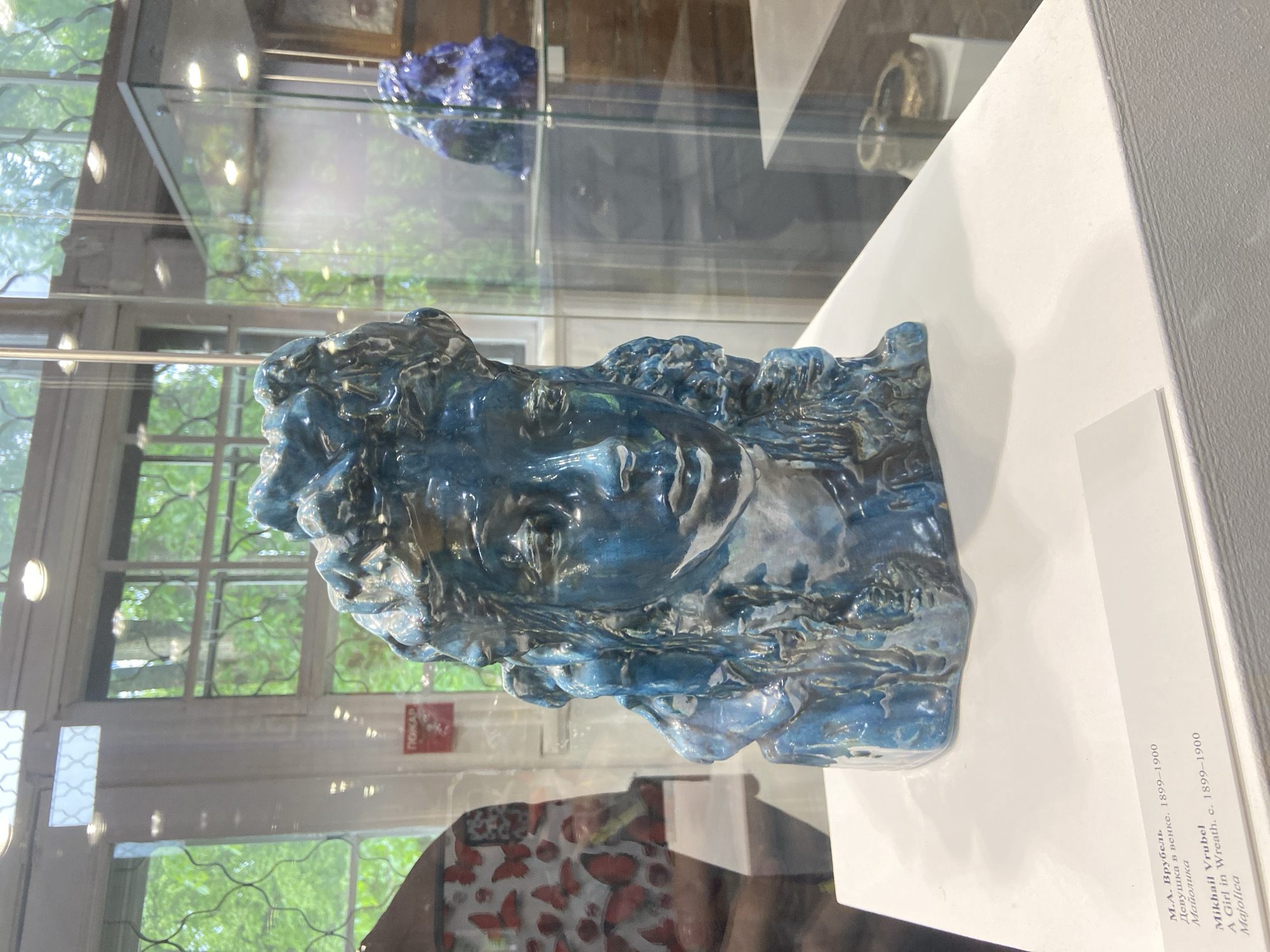 The image shows a blue glazed ceramic sculpture of a woman's head with a wreath in a glass display case. The sculpture is identified in the text as "A Girl in Wreath" by Mikhail Vrubel, c. 1899-1900. The text also identifies the location as Majolica. The scene suggests a museum or art gallery.