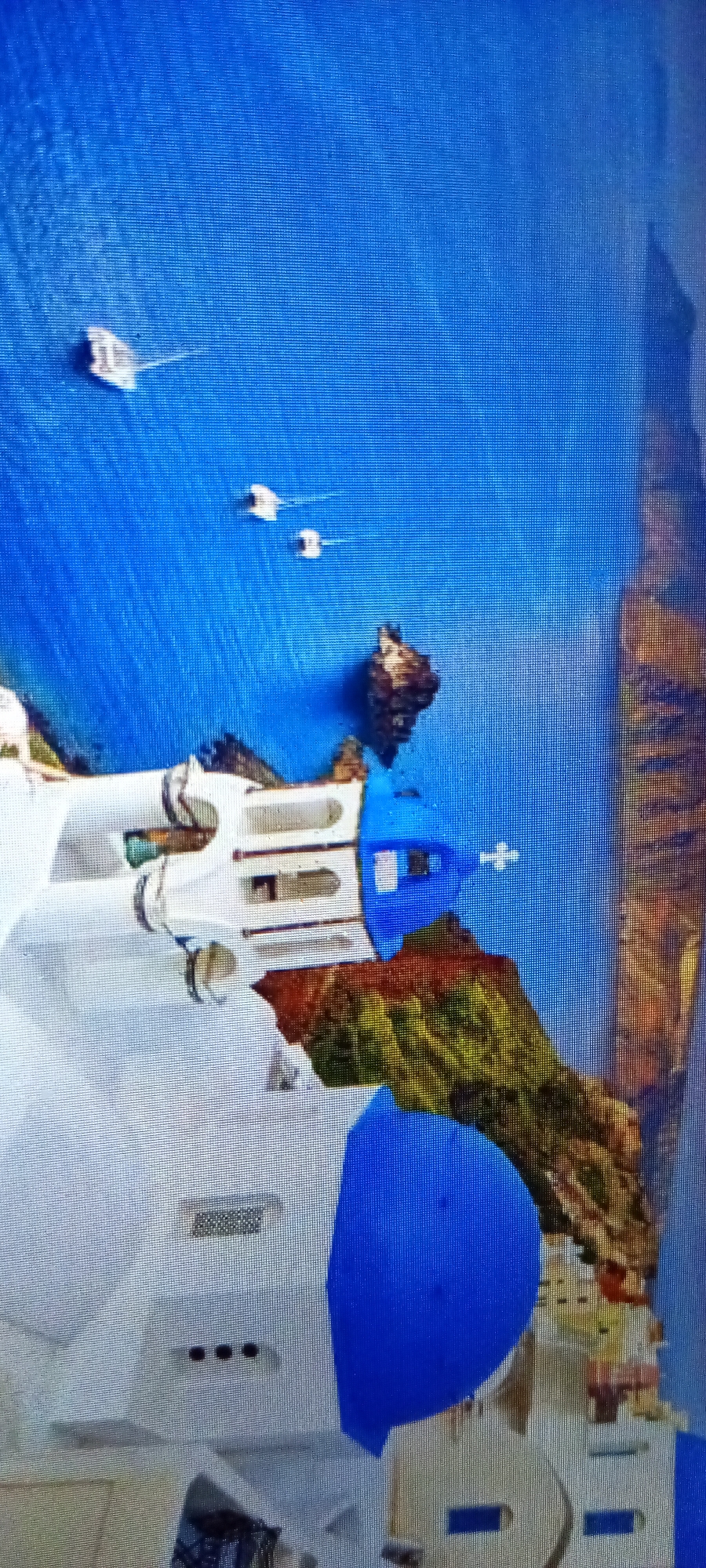 The image shows a coastal scene with white buildings featuring blue domes and accents, suggesting a Mediterranean architectural style. The structures are located near a rocky coastline with green vegetation. The sea is a deep blue, with several small boats visible in the distance. The image might be a painting or a digitally created image rather than a photograph. The prompt mentioned Lhokseumawe, Indonesia, however the architectural style in the image doesn't look consistent with the common Indonesian architecture.