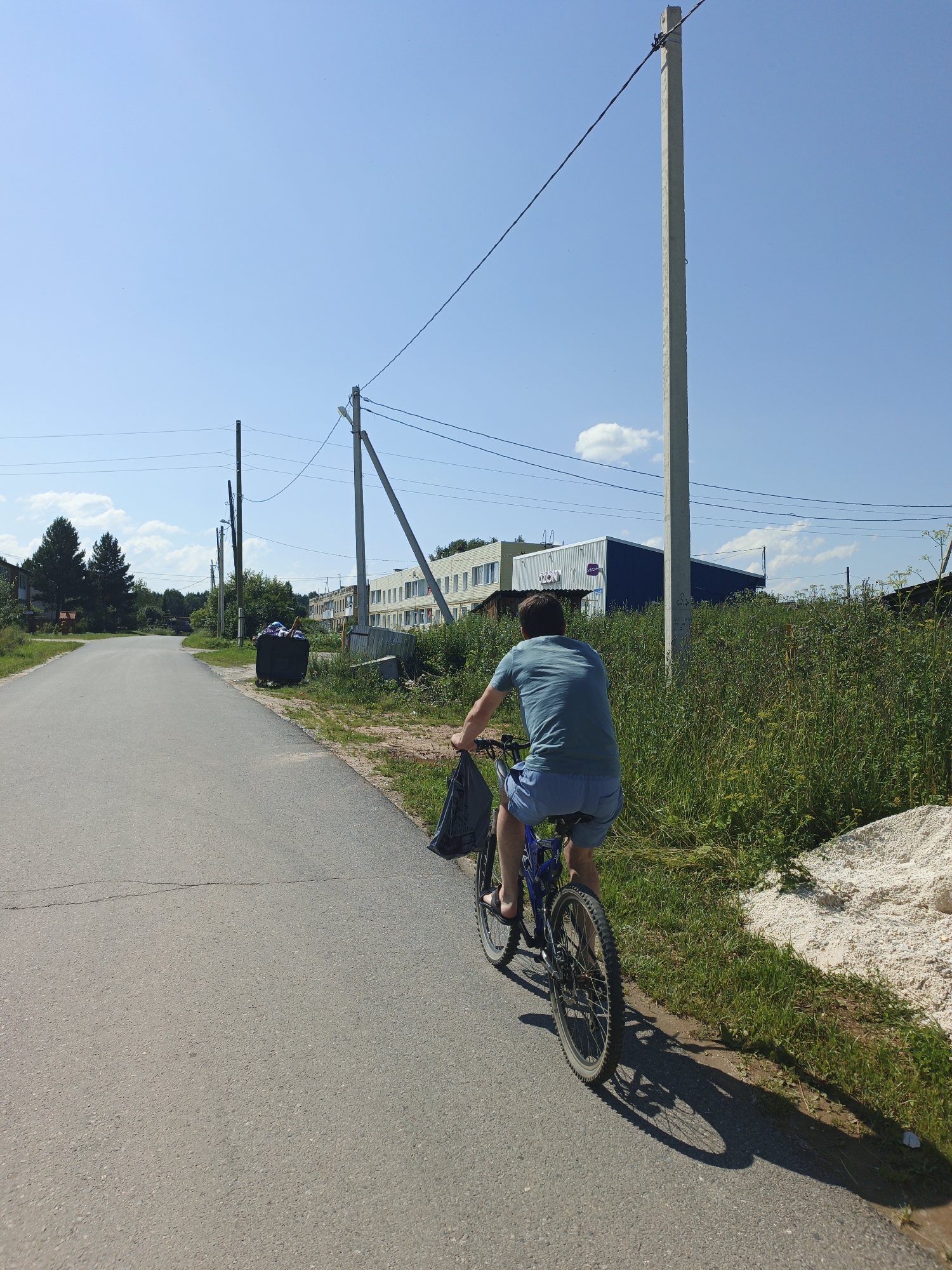 In the village of Medvedyovka, Russia, a person rides a bicycle down a paved road on a sunny day. The person is wearing a blue t-shirt and blue shorts, and is carrying a dark bag over their left shoulder. The bicycle is blue and black. To the right of the road, there is a large black dumpster filled with items, and beyond that, a tall concrete utility pole with overhead wires. Further down the road, there are buildings, trees, and dense green vegetation. The sky is clear blue with a few white clouds. A long shadow is cast by the person and bicycle onto the road.