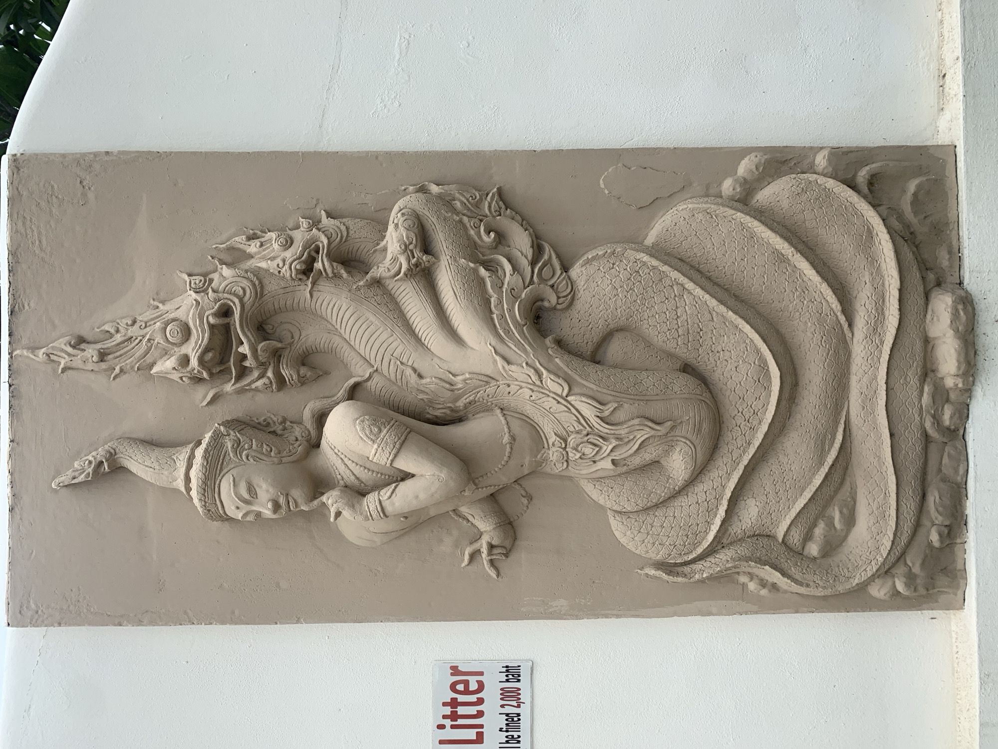 A detailed, beige-toned relief carving is affixed to a white wall. The carving depicts a figure reclining beneath the protection of a multi-headed serpent. The serpent's heads are elaborately detailed, with ornamental features. The figure is clad in stylized attire. The serpent's body curves around the figure. Below the carving, a sign warns "Litter will be fined 2,000 baht." The location is Baan Bo Phut, Thailand.
