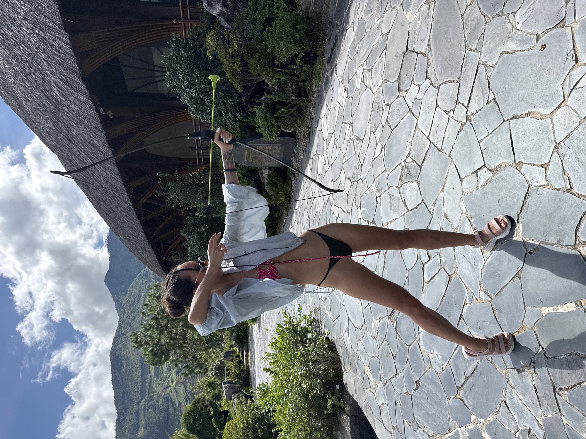 A woman is captured in an outdoor setting, posed in an archery stance, holding a black recurve bow with a yellow-green arrow nocked. She is seen from a side-back angle, with her dark hair tied in a bun and sunglasses resting on her head. Her attire consists of a light-colored, possibly white or light blue, long-sleeved shirt worn open over a pink patterned bikini top and black bikini bottoms. She is wearing light pink sandals with white soles. Tattoos are visible on her right forearm.
The scene takes place on a pathway paved with irregular gray flagstones. In the background, to the left, is a building with a distinctive dark, possibly thatched or natural fiber roof and wooden architectural details. Lush green foliage, including bushes and trees, dots the landscape around the path and the building. Further in the background, a majestic range of green, forested mountains stretches under a clear blue sky dotted with white clouds, suggesting a bright daytime setting. A stone slab with unidentifiable East Asian script is partially visible behind the bow. The overall environment, consistent with Púng Luông, Vietnam, suggests a scenic resort or natural park setting.