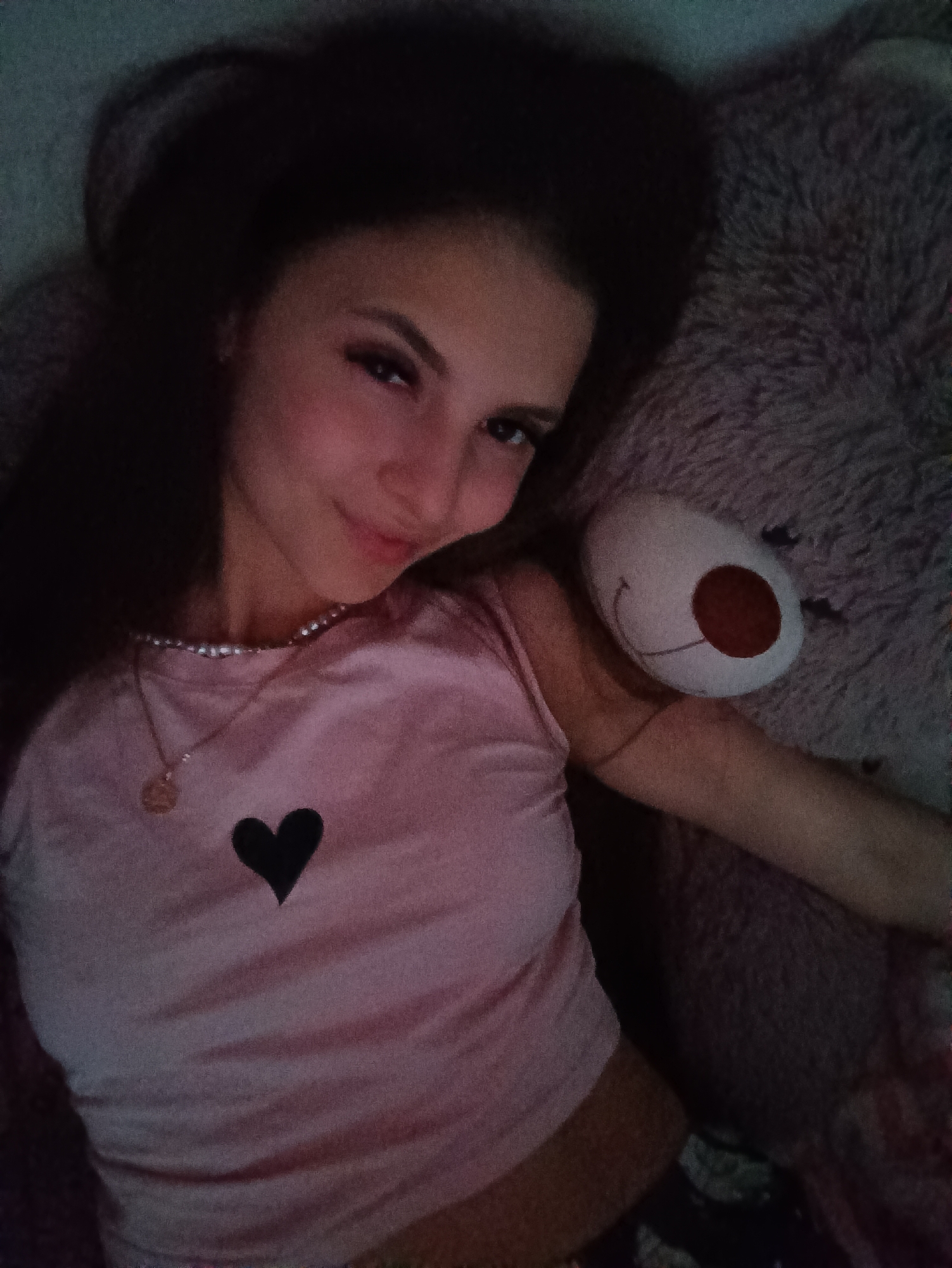 A young woman lies on a fluffy, grey surface next to a stuffed animal. She is wearing a light pink shirt with a black heart on it, a beaded necklace, and a pendant necklace. Her long brown hair is spread around her head, and she is looking towards the camera with a slight smile. The background is dark and indistinct. The city and location are unknown.