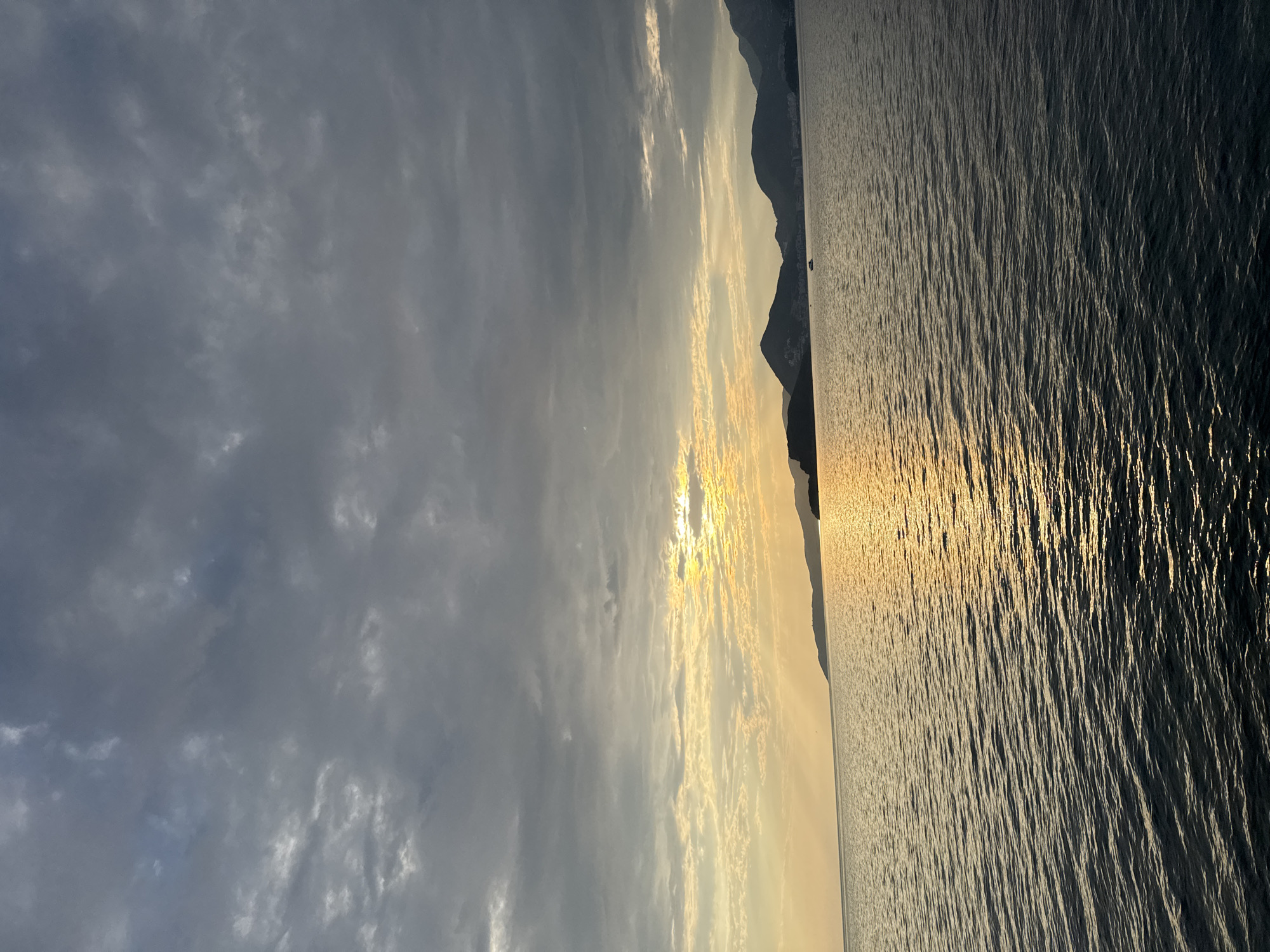 A wide shot captures a serene seascape at sunset near Sveti Stefan, Montenegro. The sky is a dramatic canvas of heavy, gray clouds, with a sliver of golden light breaking through near the horizon, casting a shimmering path across the rippling, dark water. Silhouetted against the luminous sky are the rugged, verdant outlines of mountains that form the coastline, with faint structures of buildings visible along their slopes. A small, dark boat is a solitary object on the water, adding a sense of scale to the vastness of the scene. The overall impression is one of tranquil twilight, with the last rays of the sun painting the sky and sea.