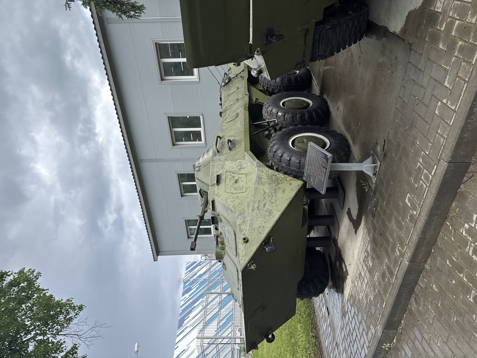 The image depicts a BTR-50PK armored personnel carrier (APC) on display in Votkinsk, Russia. The APC is painted in a faded green camouflage pattern. It is mounted on a metal stand, suggesting it is a museum piece or static display. The APC's tires are large and black with white rims. A small sign with text in Russian stands next to the vehicle. The ground is paved with gray bricks. In the background is a light gray building with windows and another building with blue and white stripes. The sky is overcast, indicating a cloudy day. A tree is visible on the left side of the image.