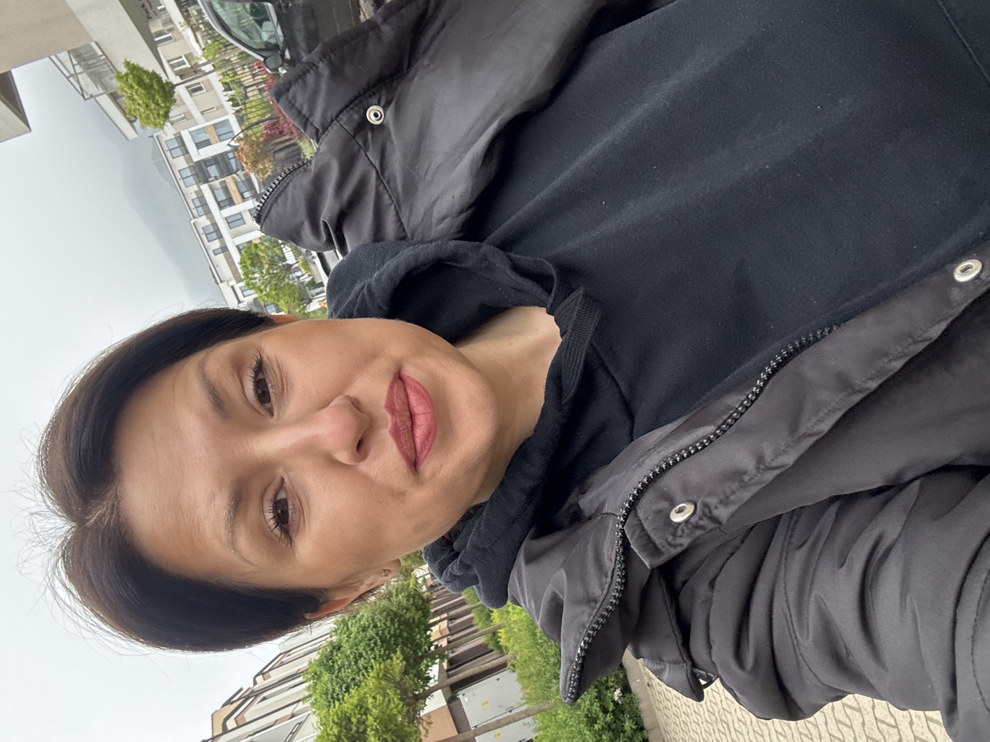 A woman in a black hooded sweatshirt and a black jacket is shown in a close-up, slightly angled portrait. She has short, dark brown hair, and brown eyes. Her lips are pink.
The background shows an urban landscape. Modern, multi-story residential buildings are visible. Green trees and bushes line the street. The sky appears overcast. The scene is likely in Sofia, Bulgaria, as indicated in the request. The image seems to be taken outdoors.
