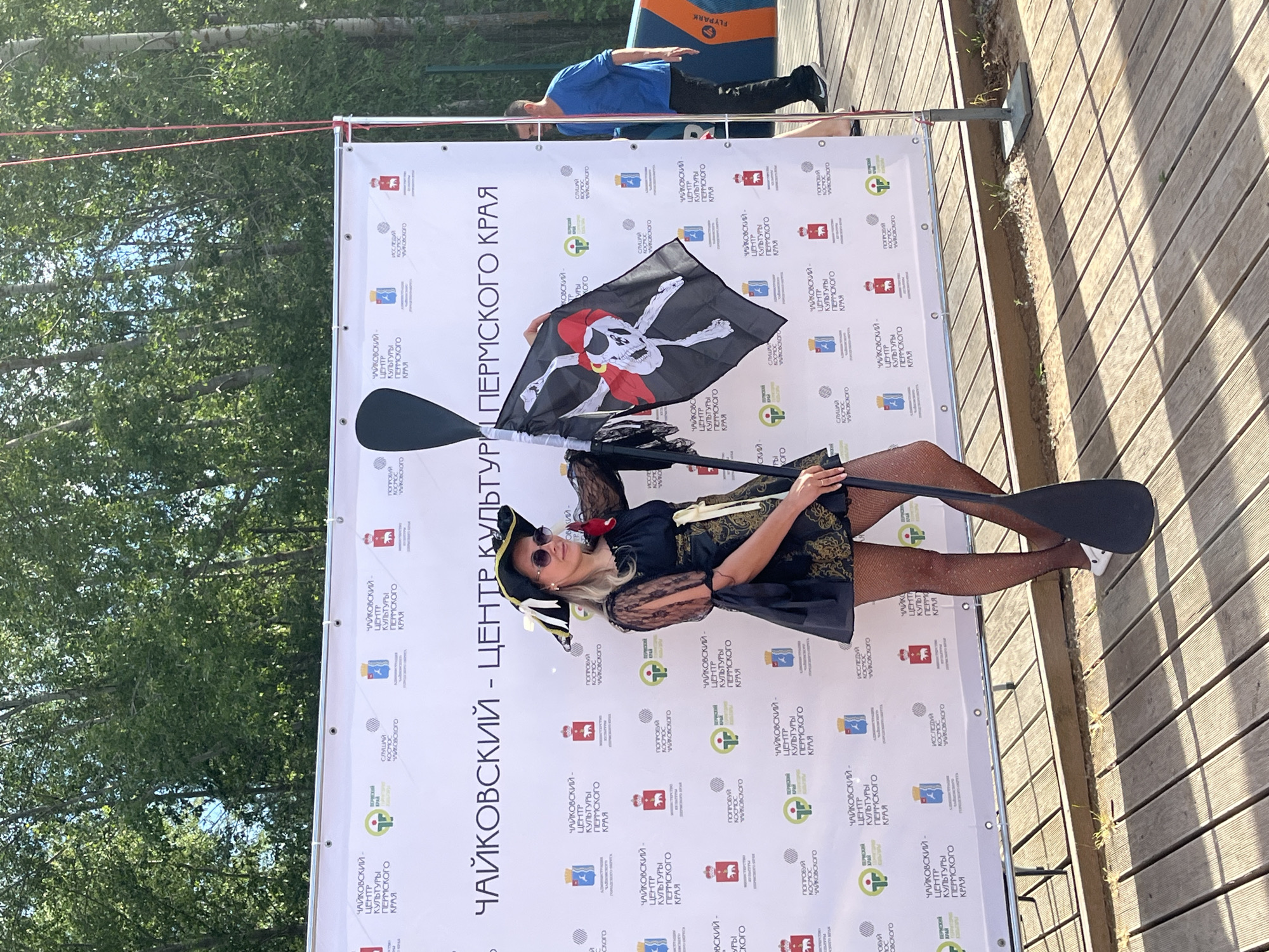 A woman in a pirate costume stands on a wooden platform in front of a banner. She is wearing a pirate hat, sunglasses, a black dress with gold embellishments, and patterned tights. She holds a pirate flag and a paddle. The banner behind her displays logos and text in Russian. A man in a blue shirt and black pants walks in the background, and an inflatable paddleboard is visible to the right. The scene appears to be outdoors, with trees in the background. The text on the banner mentions "Чайковский - Центр культуры Пермского края," which translates to "Chaikovsky - Center of Culture of Perm Krai," indicating the location is in the city of Chaikovsky, Russia.