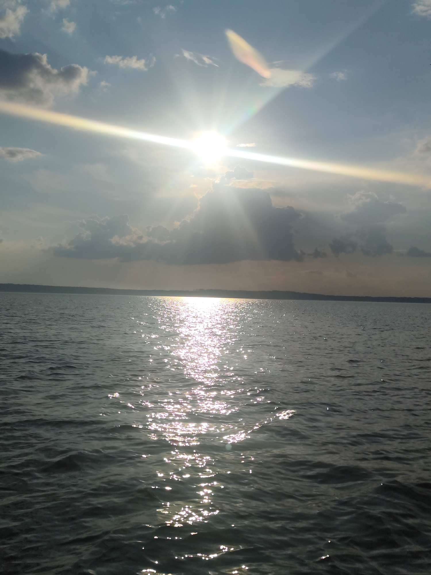 The image depicts a vast body of water, likely a lake or sea, under a cloudy sky illuminated by the sun. Sunbeams pierce through the clouds, creating bright streaks of light that reflect off the rippling surface of the water, forming a shimmering path. The horizon line is barely visible, suggesting a wide expanse of water. The water itself appears dark and slightly choppy. The sky is a mix of blue and grey clouds, with the sun positioned high in the frame. No people or man-made structures are visible in the image. The location of Галево, Russia is not discernible from the visual information provided in the image.