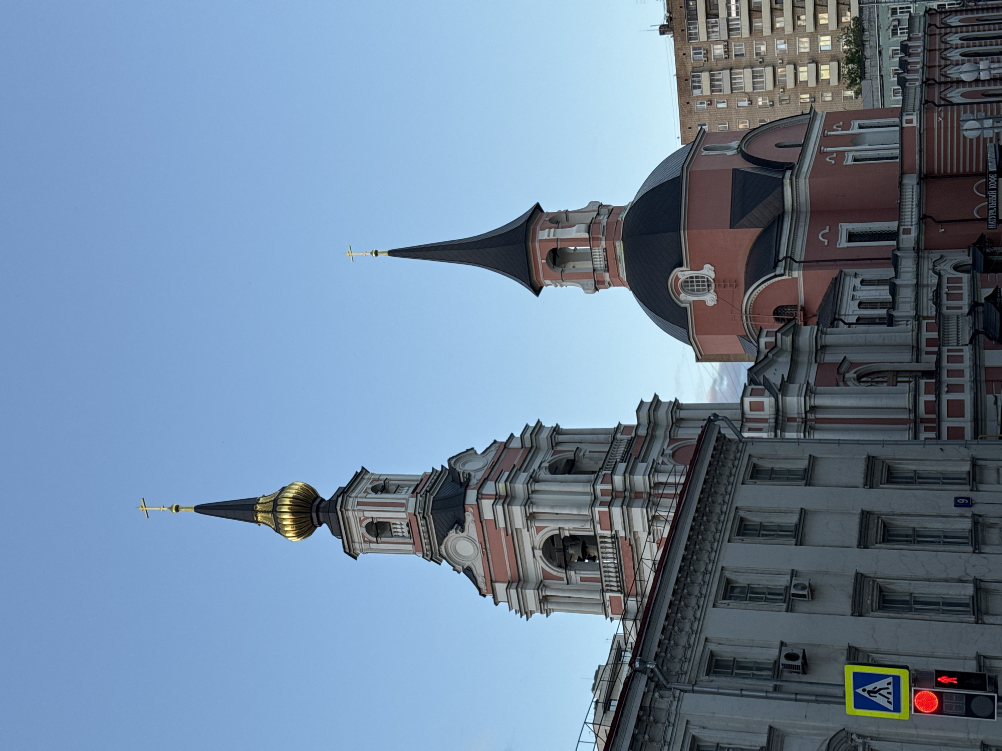 The image shows a view of two Russian Orthodox churches in Moscow, Russia, with a clear blue sky. The church on the left has a tall bell tower with a golden-domed spire topped by a cross. The tower is white with pink accents and features multiple levels with arches and decorative columns. Below the bell tower, a section of a grey building with many windows and air conditioning units is visible.
The church on the right features a pink structure with a dark, rounded dome and a slender, black spire topped with a cross. This church also has decorative white elements and arched windows. In the background, a large, multi-story apartment building with numerous windows is visible, along with some trees and a smaller green building.
In the foreground, on the left side of the image, a pedestrian crossing sign with a blue background and a white silhouette of a walking person is displayed. Below it, a traffic light shows a red signal for pedestrians, indicating "Don't Walk." A street sign with the number "9" is also visible. The overall scene depicts an urban environment with historical religious architecture juxtaposed with modern residential buildings.