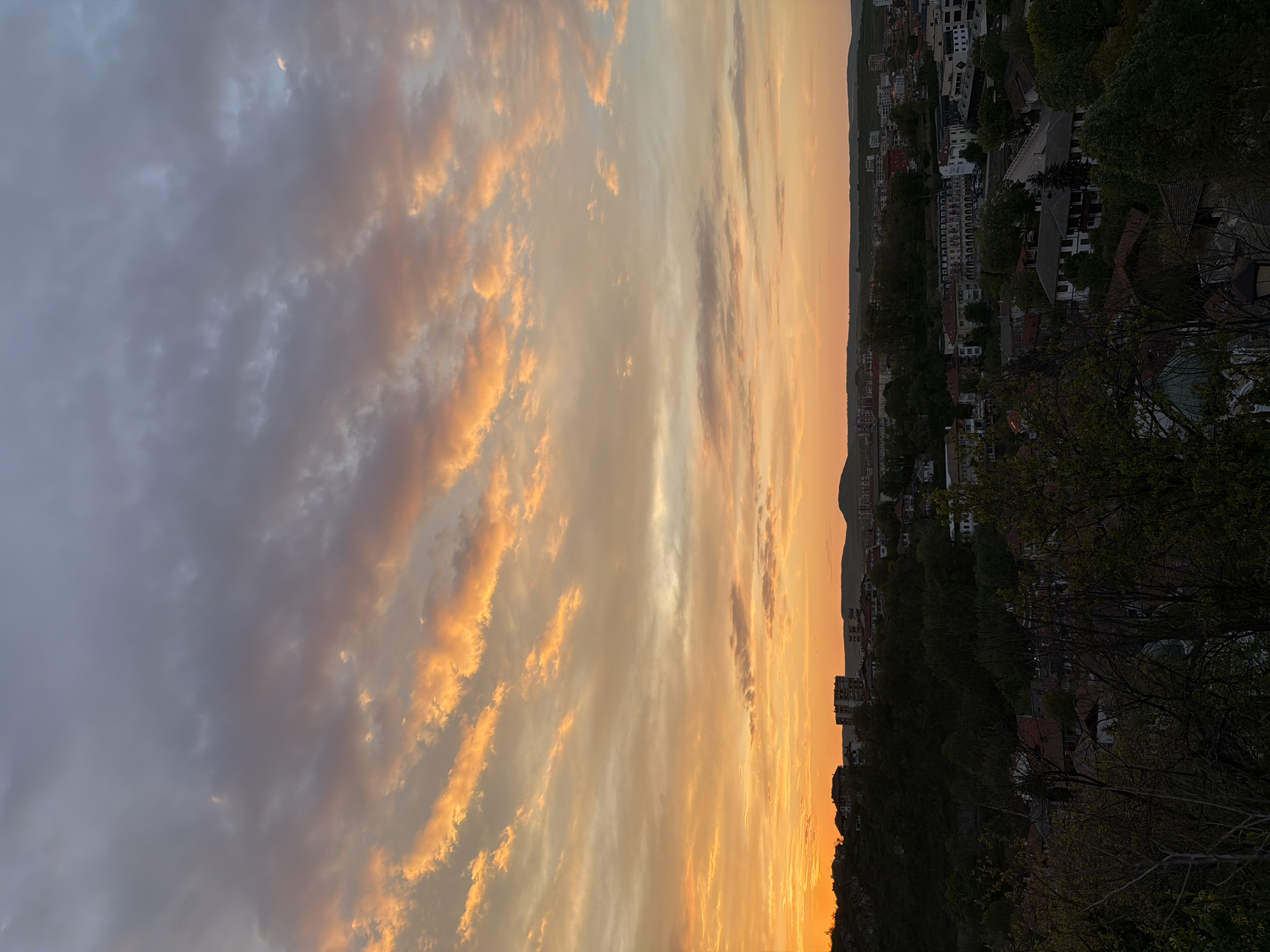 The image shows a landscape view of Lovech, Bulgaria, during a vibrant sunset. The sky is filled with a mix of orange and gray clouds. Below the sky, a mountain can be seen with the silhouette of a village. The foreground is dominated by trees. The image is taken from a slightly elevated position, offering a broad view of the setting.