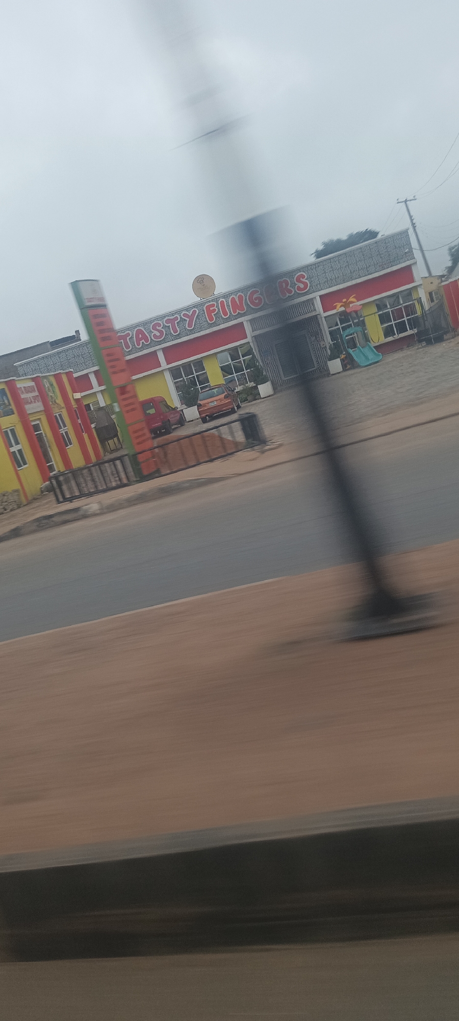 The image presents a motion-blurred, oblique view of a roadside commercial establishment, likely a restaurant, in Jos, Nigeria, taken from a moving vehicle. The primary subject is a brightly colored building featuring prominent yellow walls and red horizontal trim, with a grey textured upper facade and large windows. Above the entrance, a large sign clearly displays the name "TASTY FINGERS" in red capital letters with white outlines. A circular emblem is also visible on the sign.
In front of the building, several vehicles are parked, including a red car and an orange or brown car. A blue and white outdoor slide is visible on the right side, suggesting a play area. To the left of the main building, a smaller, similarly styled yellow and red striped structure is partially visible. A tall, vertical signpost, mainly green and red, stands near this smaller structure but is heavily blurred.
The foreground consists of a wide, reddish-brown dirt shoulder adjacent to a grey asphalt road, both exhibiting significant motion blur. A dark, heavily blurred utility pole extends vertically from the bottom to the top right quadrant of the image. The sky is overcast, indicating a cloudy daytime.