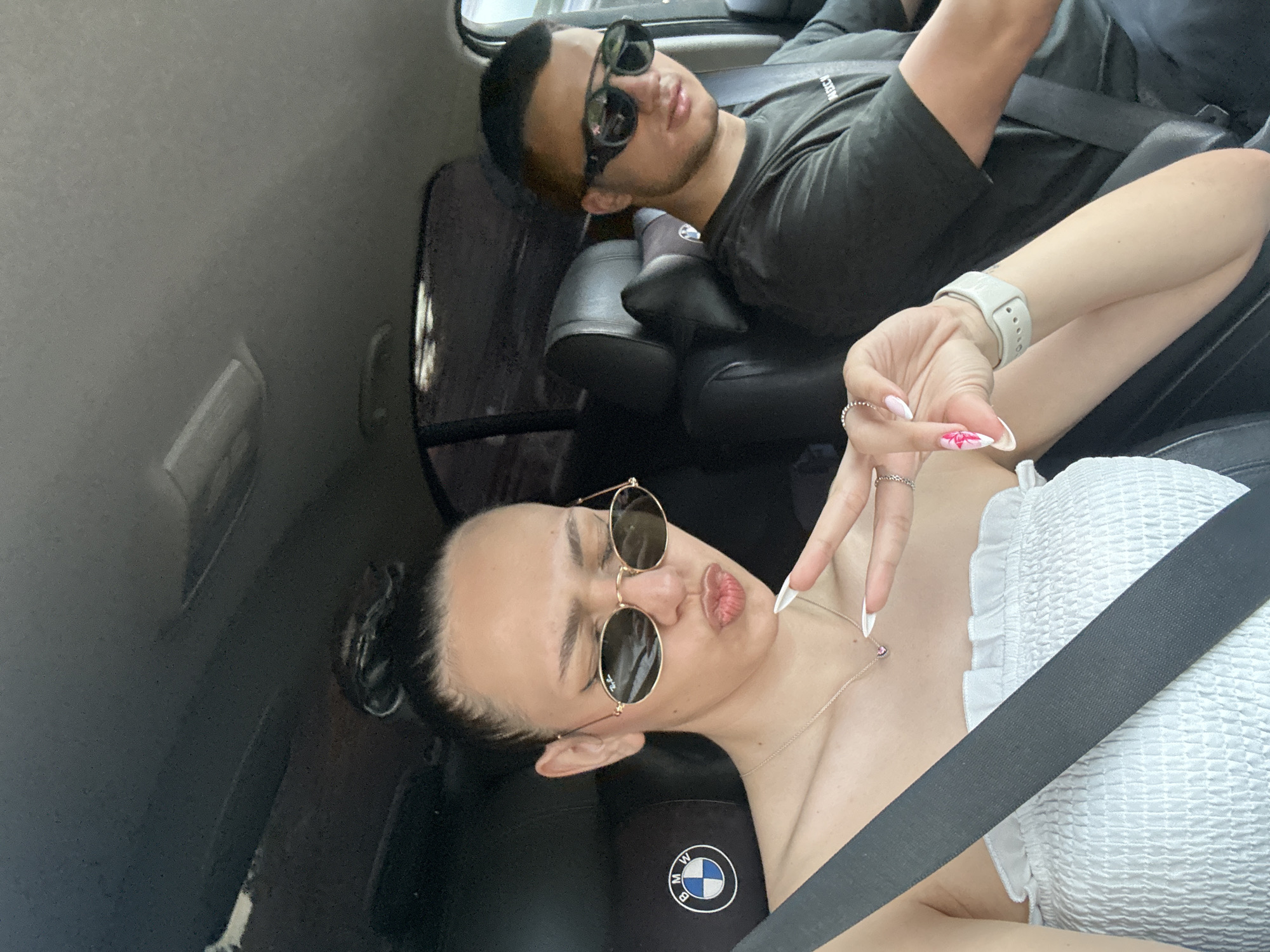 Two individuals are sitting in the back seats of a BMW. The person in the foreground is wearing a white top and sunglasses, and is making a gesture with their hand. The person in the background is wearing a dark t-shirt and sunglasses. The interior of the car is black leather. There is no evidence to suggest that the photo was taken in Borovo, Bulgaria.