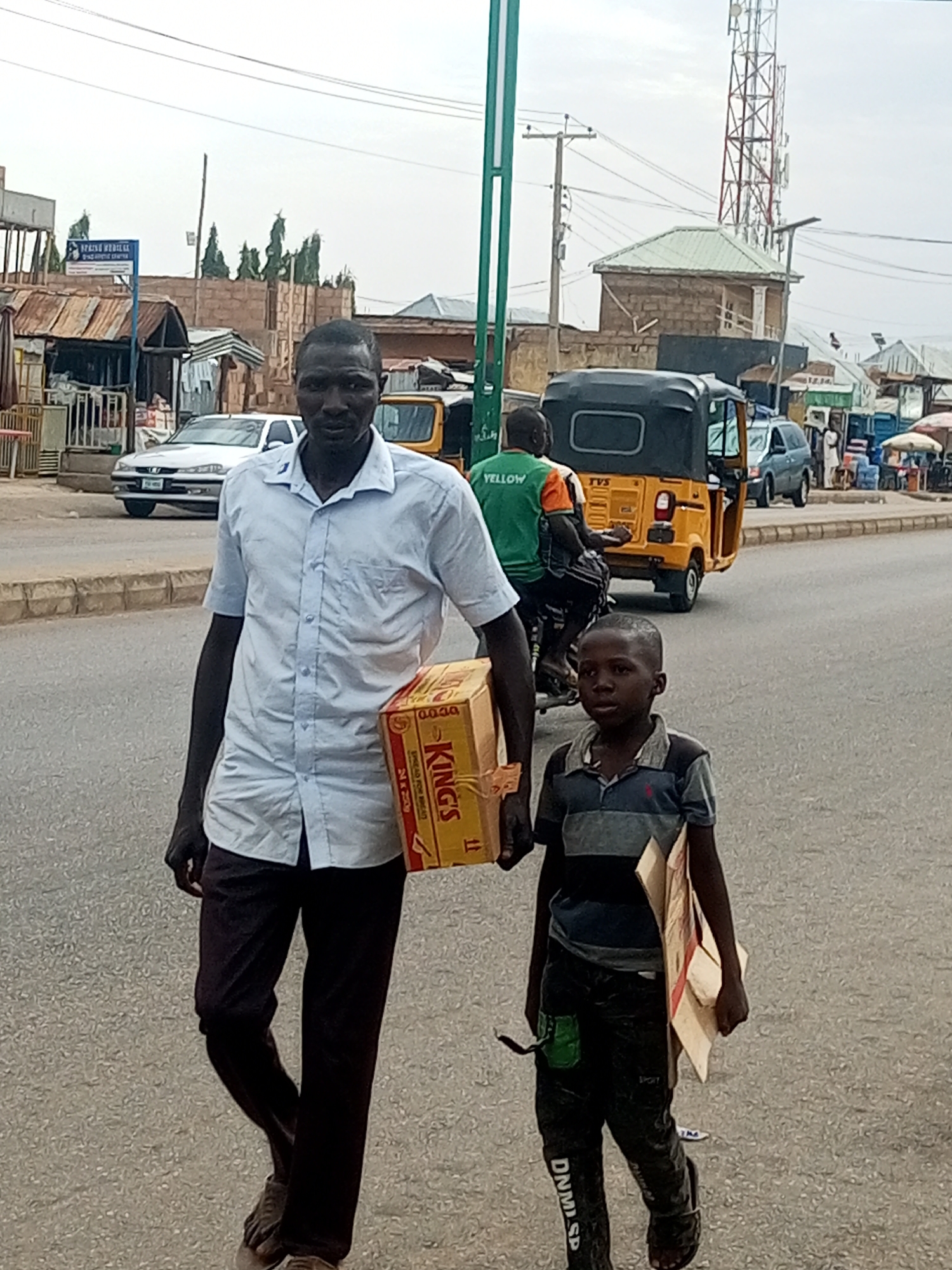 In Tempure, Nigeria, a man and a young boy walk down a paved street. The man, in a light blue short-sleeved shirt and dark trousers, carries a large cardboard box labeled "KING'S". The boy, wearing a striped t-shirt and dark pants with "DNM SP" printed on them, carries a smaller cardboard box.
The background reveals a bustling street scene. To the left, market stalls with corrugated metal roofs line the road, and a white car is parked. Further down the street, a yellow auto-rickshaw, possibly a "TVS" model, is visible, with a man in a green vest riding on it. A cell tower stands tall in the distance.
The sky is overcast, suggesting a cloudy day. The overall impression is of everyday life in a Nigerian town, with people engaged in carrying goods along a busy thoroughfare. The man's expression is neutral, and the boy's gaze is directed forward. The scene captures a moment of transit, with both individuals focused on their journey. The visible text on the box identifies the brand as "KING'S" and notes "24 x 250g," indicating the contents are likely beverages or food items. The "SPRING VEDILAL" text on a sign suggests a nearby establishment.