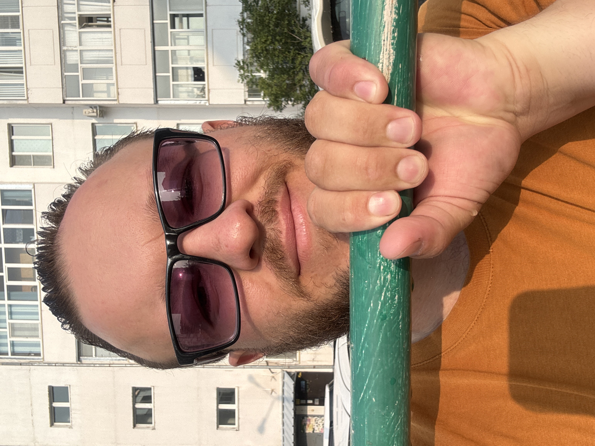 A close-up, eye-level, outdoor, selfie-style shot shows a man with dark, slicked-back hair, wearing black-rimmed sunglasses with pink lenses, and an orange t-shirt. He is holding onto a green, wooden bar with his right hand. The man has a beard and stubble. In the background, a pale, multi-story apartment building with numerous windows and balconies is visible. The time of day appears to be daytime, with sunlight casting shadows. The man's face is partially obscured by his hand and the bar, and his gaze is directed towards the camera. The overall setting suggests an urban environment, possibly a park or recreational area within Blagoveshchensk, Russia, given the architectural style of the building.