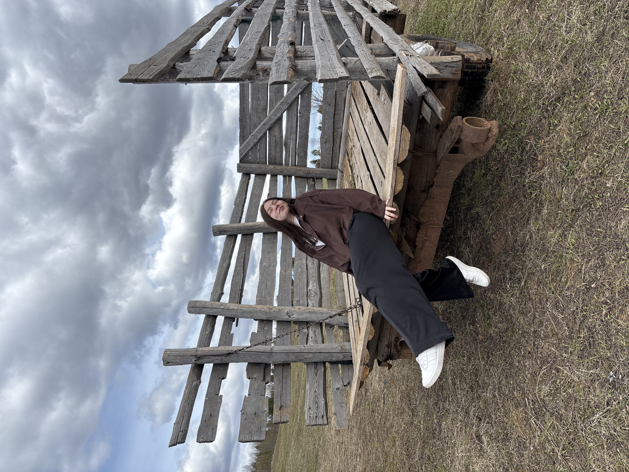 The image shows a young woman standing next to an old, wooden farm wagon on a grassy field under a cloudy sky. The wagon is constructed of rough-hewn logs and planks, with a high, slatted frame above the bed. The woman is wearing a brown jacket, black pants, and white sneakers. She is leaning against the wagon with her eyes closed. The location is most likely somewhere near Петропавловск, Russia.