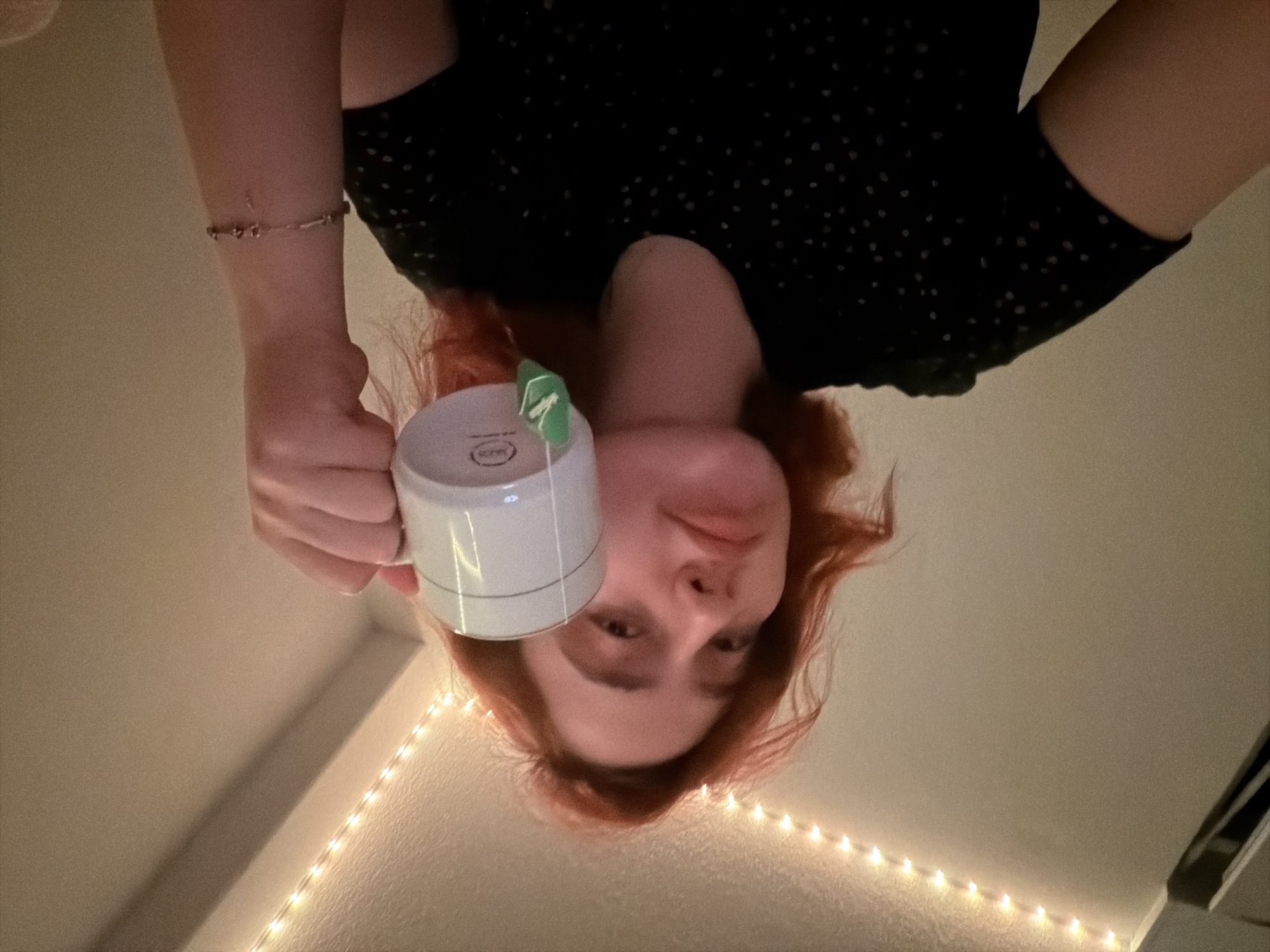 A person with reddish-orange hair is holding a white mug with a tea bag, looking towards the camera. The person is wearing a black shirt with white polka dots. A bracelet is visible on their right wrist. The background is a plain wall illuminated by a string of warm-toned lights along the ceiling. There is no indication of the location being Strasbourg, France.