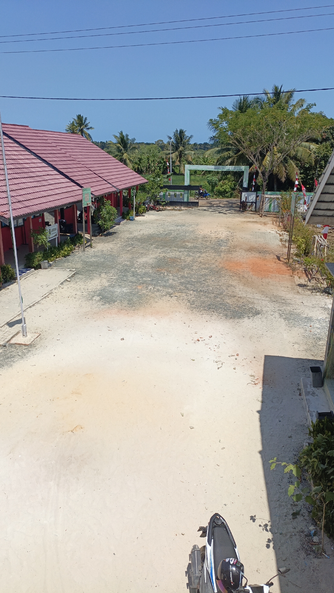 The image depicts a schoolyard in Saring Sungai Binjai, Indonesia, on a clear, sunny day. The foreground is dominated by a sandy, somewhat uneven ground, with patches of darker soil and some scattered red-orange dust. A portion of a motorcycle, its seat and rear fender visible, is parked in the lower right corner.
To the left, a building with a red tiled roof extends into the frame. It appears to be a school building, with visible support columns and a white pole, possibly a flagpole, in front. Some greenery, including trees and bushes, is present along the building's edge and further into the yard.
In the background, a green archway marks an entrance or passage, with a black gate beneath it. Beyond the archway, there are more trees, including palm trees, and dense vegetation suggesting a rural or semi-rural setting. Indonesian flags, red and white, are visible near the entrance. Two individuals are seen near the gate, one on a motorcycle. The sky is a clear, bright blue.