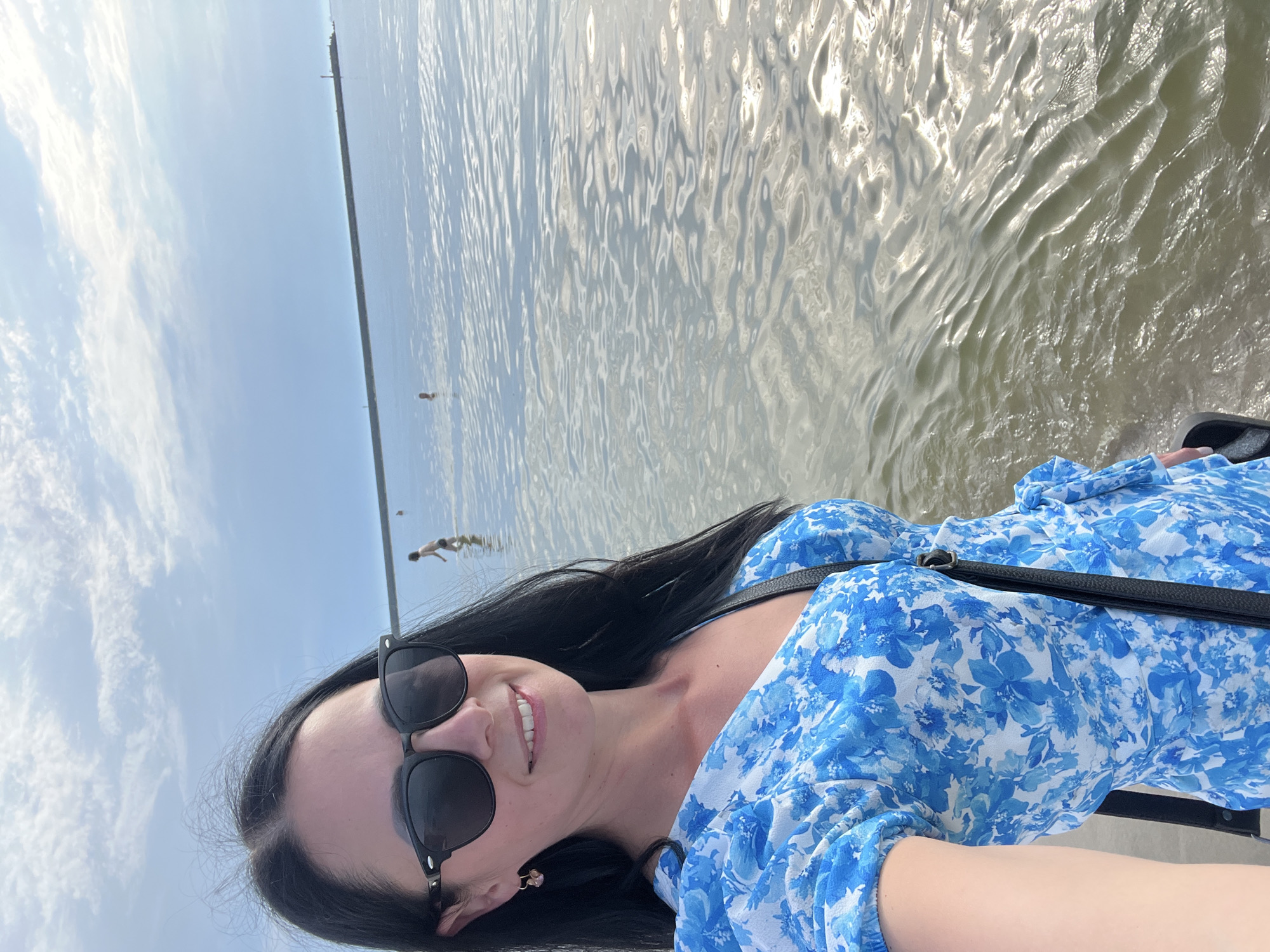 A woman with dark hair, wearing sunglasses and a blue floral dress, takes a selfie at the water's edge in Baltiysk, Russia. She stands on a sandy or rocky shore, with calm water stretching out before her. In the distance, a long stone breakwater extends into the sea, with a lighthouse at its end. Several people are visible in the water, wading or swimming. The sky is partly cloudy, with a bright sun reflecting off the water's surface. The overall scene suggests a pleasant day at the beach.