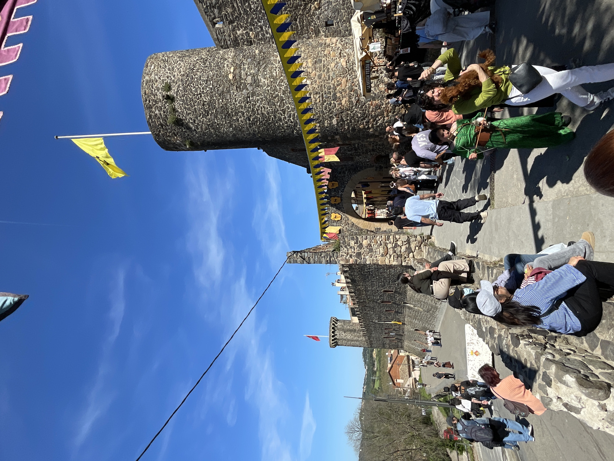 The image depicts a scene in Hostalric, Spain, likely at a festival or fair near the medieval castle. In the foreground, people are sitting along a stone wall, seemingly enjoying the event. The main focus is the stone castle, with banners and flags hanging from it. Several people are gathered near the castle, likely vendors or attendees of the event. The sky is clear and blue. The image captures a bustling atmosphere with the castle as a historical backdrop.