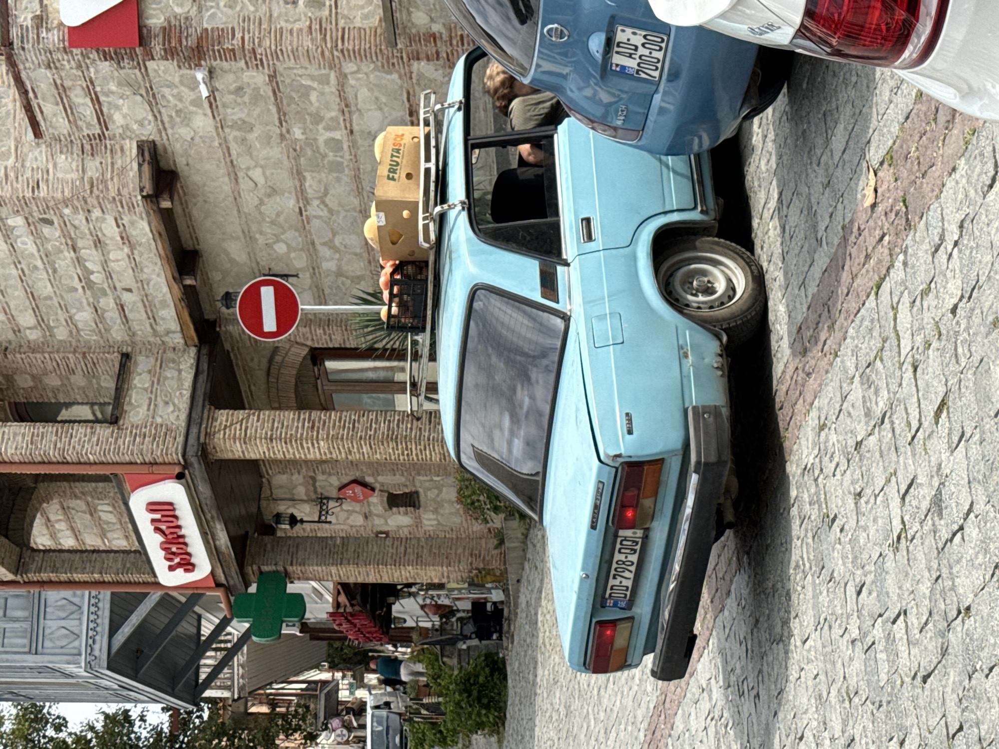 A light blue Lada car is parked on a cobblestone street in Sighnaghi, Georgia. The car has a roof rack with a black crate of fruit and a cardboard box labeled "FRUTASOL" on top. The license plate of the Lada reads "DD-798-QQ". To the right of the Lada, a blue Fiat car is parked, with its license plate reading "AD-700-00".
In the background, a brick building features a red and white sign with Georgian script, and a green cross sign. A red and white circular "no entry" sign is also visible. A person is partially visible in the driver's seat of the Lada, leaning back. Other people and vehicles are present further down the street. The environment appears to be a commercial or residential area, with shops and buildings lining the street.