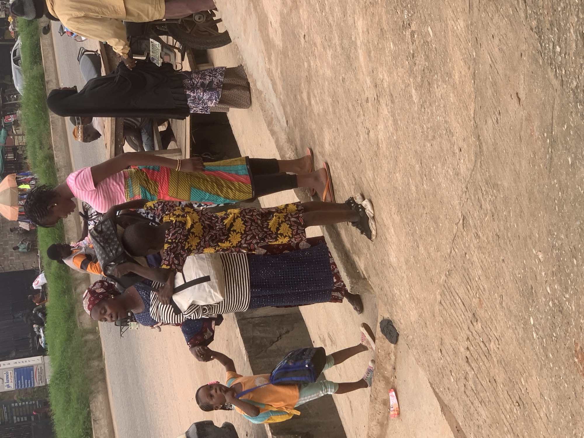 The image depicts a street scene in Shagamu, Nigeria. In the foreground, a young girl wearing an orange shirt and light blue leggings carries a blue backpack and another bag. She is walking on a concrete surface alongside a woman in a blue and white striped top and a dark blue skirt, who is carrying a white and black bag. To the right, another woman in a black hijab and a patterned skirt stands near a motorcycle. In the background, several other individuals are visible near a market stall with various goods. A road with a parked car and a motorcycle is also present. The environment appears to be a bustling urban area with a mix of people and activities.