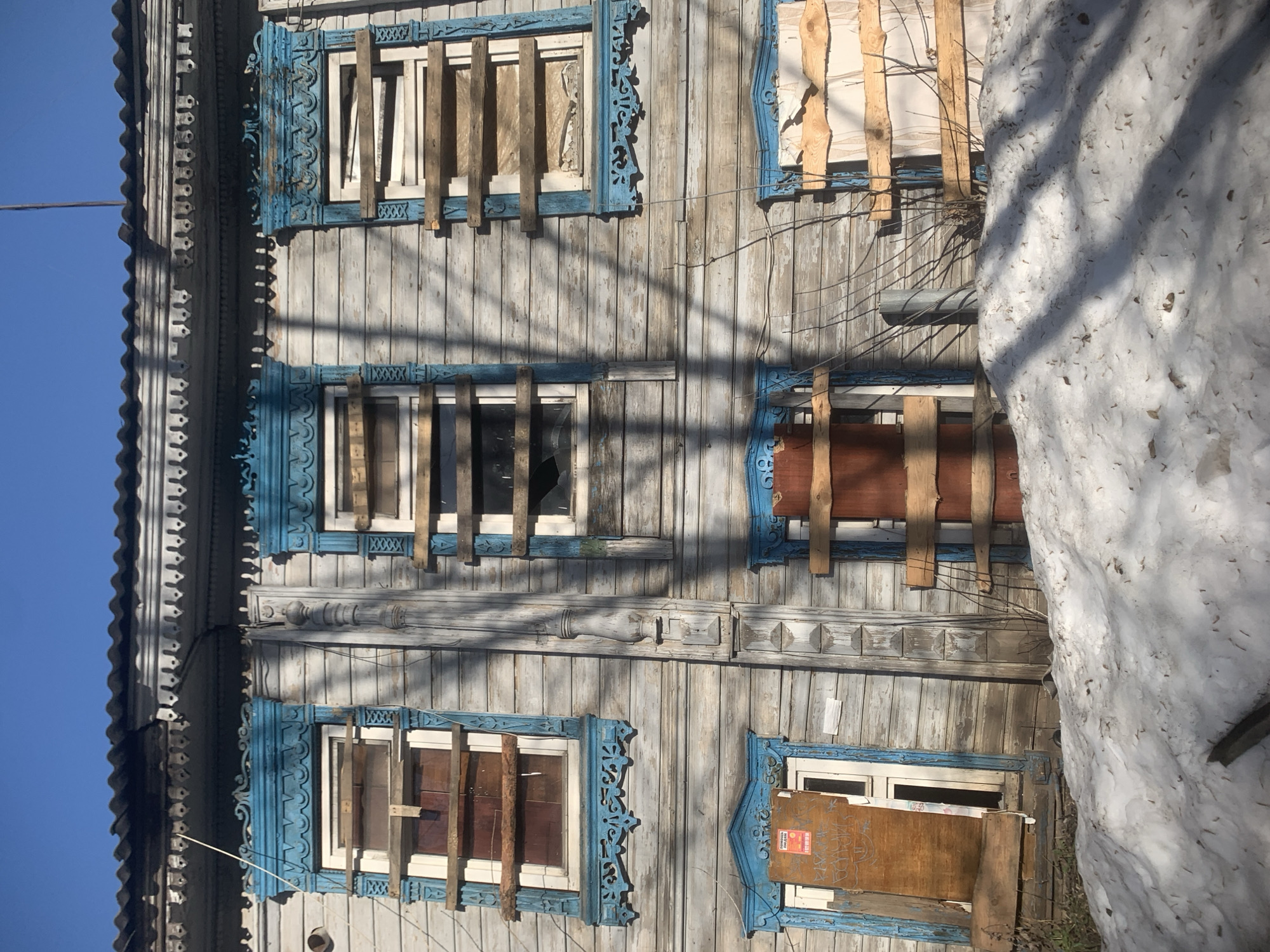 The image shows the facade of a weathered, two-story wooden building, likely located in Izhevsk, Russia, based on the architectural style. The building is constructed with vertical wooden planks, exhibiting a faded gray-blue paint. The windows are boarded up with wooden planks in varying states of repair, and the window frames are painted a bright blue with decorative carvings. One window has broken glass. A large pile of snow is visible on the right side of the image, partially obscuring the lower part of the building. Bare tree branches cast shadows on the facade. The sky is clear and blue in the upper left corner. A wire runs horizontally along the top of the building.