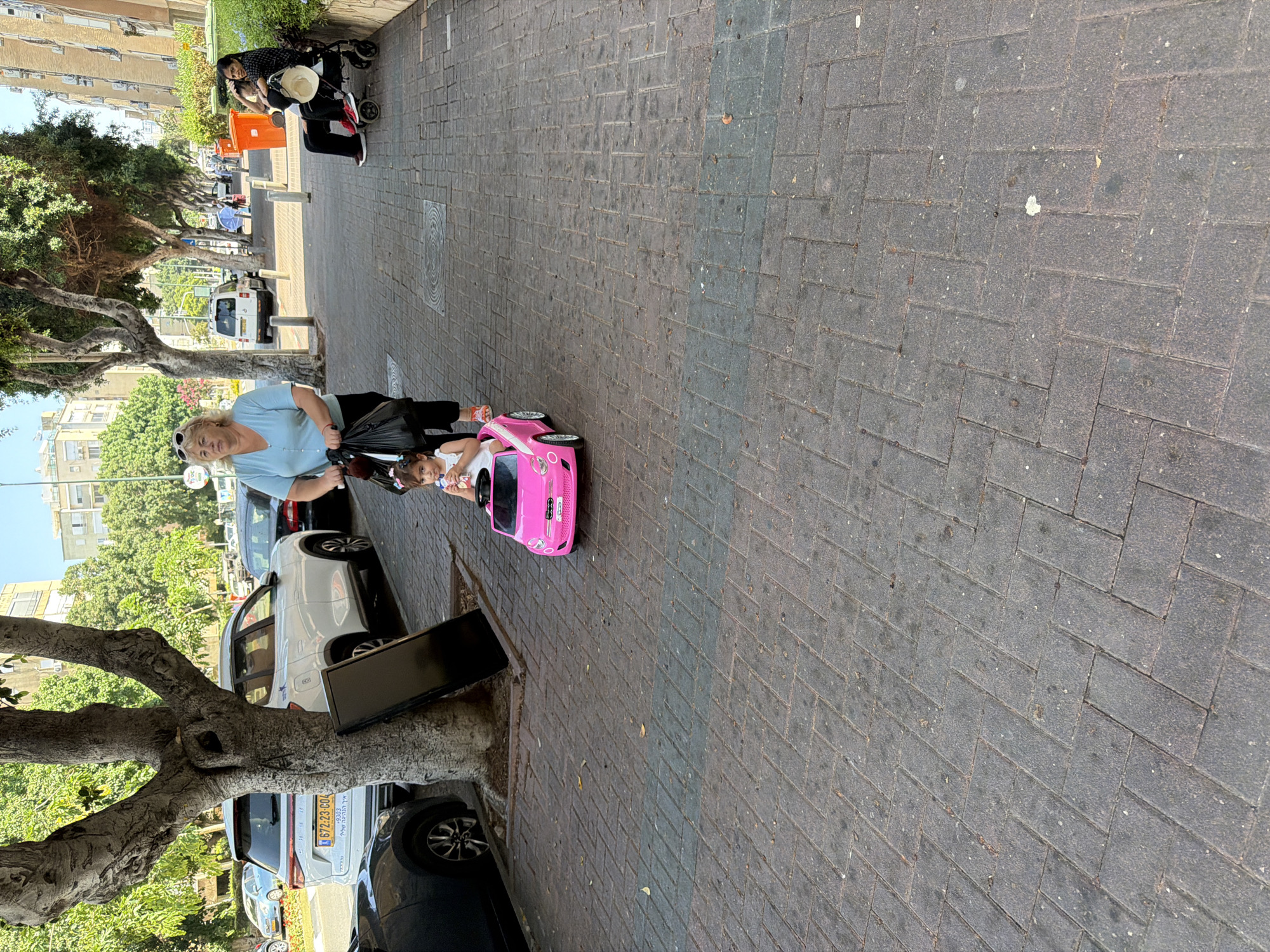 A woman with blonde hair wearing a light blue t-shirt and sunglasses is pushing a pink toy car with a young child inside. The child is wearing a white top and has dark hair. They are on a paved sidewalk lined with trees. In the background, there are parked cars and buildings. Another person, dressed in dark clothing, is with a stroller further down the sidewalk. The scene appears to be outdoors on a sunny day. The image is taken from a slightly elevated perspective. Some visible text on a license plate reads "672.23 COL".