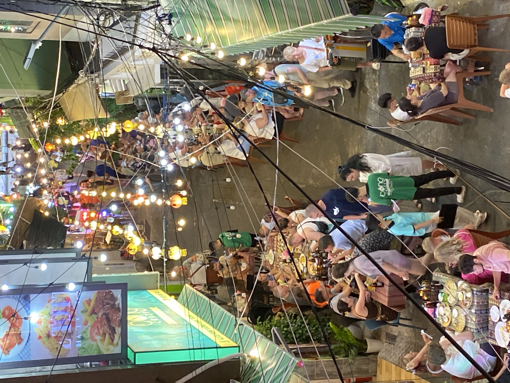 The image depicts a busy street scene in Nha Trang Ward, Vietnam, likely during the evening given the abundance of string lights illuminating the area. The street is lined with restaurants and shops, some with outdoor seating where patrons are dining. Numerous people are visible, some seated at tables, others walking or standing. Many of the diners appear to be tourists, judging by their clothing and demeanor. The atmosphere is lively and festive, with an emphasis on communal dining and socializing. The architecture suggests a bustling urban environment, with buildings on either side of the narrow street, adorned with awnings and signage. The presence of many overhead wires indicates a well-developed but potentially cluttered urban infrastructure.