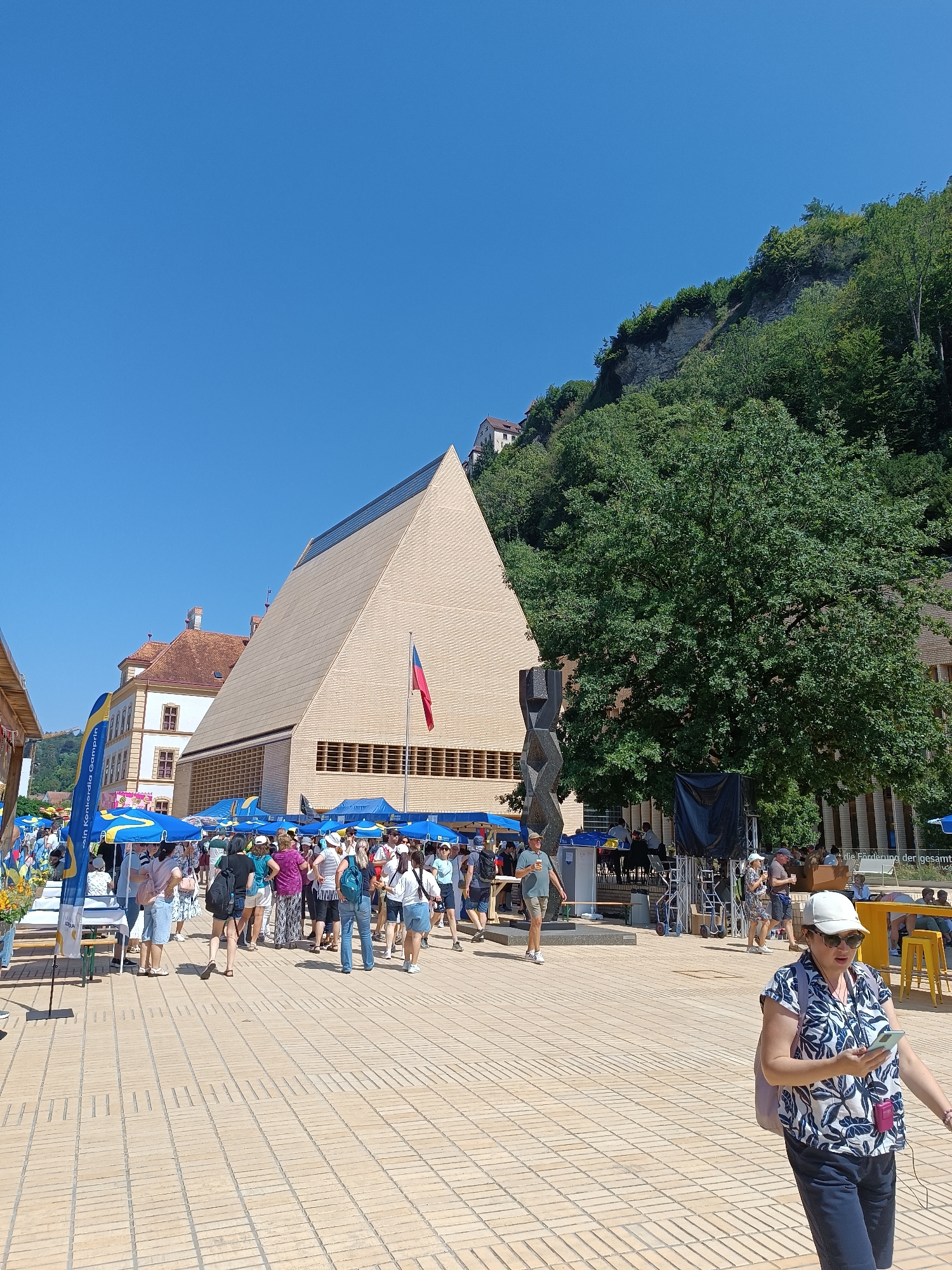 This is a wide-angle, eye-level outdoor shot of a busy pedestrian area in Vaduz, Liechtenstein. The foreground is a paved plaza made of light-colored bricks. In the mid-ground, a large, modern, beige, pyramid-shaped building with a dark-tinted sloped roof dominates the scene. In front of it, a crowd of people are gathered, many standing under blue tents and umbrellas. A flag is flying from a pole in front of the pyramid building. To the left of the pyramid building, an older, ornate building with a reddish-tiled roof is visible. A large, dark green tree stands to the right of the pyramid building, with more greenery and a cliff face in the background. A stage with musical equipment is set up near the tree. Several people are walking and interacting in the plaza, some looking at their phones, others conversing. The sky is clear and blue. The overall atmosphere suggests a public event or festival.