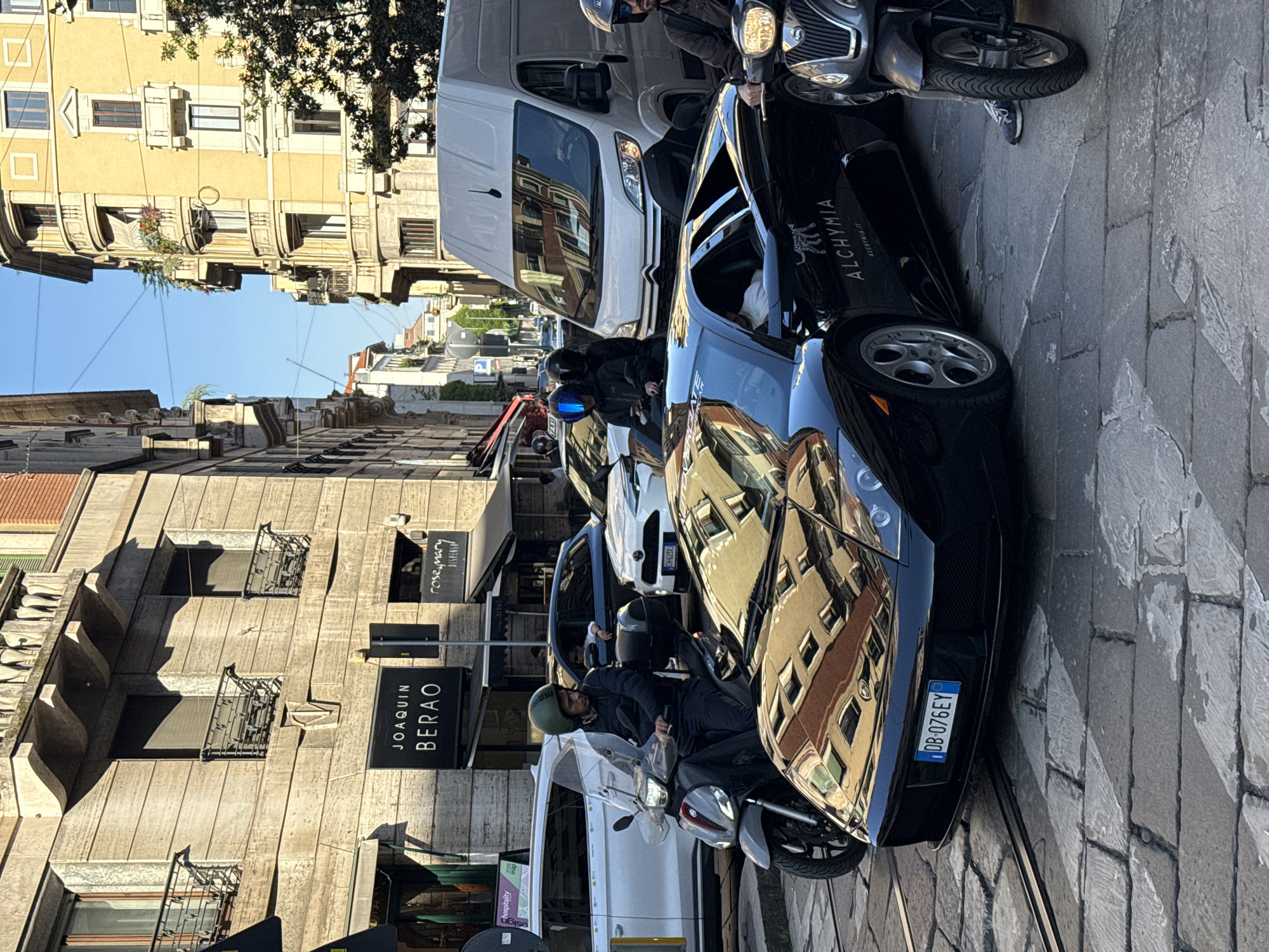 The image depicts a bustling street scene in Milan, Italy, captured on a bright, sunny day. The perspective is rotated 90 degrees clockwise, showing the sky on the right and buildings on the left.
In the foreground, a sleek black sports car is prominent, its highly reflective surface mirroring the surrounding architecture and the clear blue sky. An Italian license plate, "DB-076EY," is clearly visible on its rear. Next to and behind the sports car are several motorcycles and scooters, with riders wearing helmets, suggesting active urban traffic or parking. A silver scooter is closest to the viewer.
Further down the street, a white delivery van, identifiable as a Citroën, is present, with other vehicles including a blue car showing a partial license plate "CH-625X," and a "TAXI" sign in the background. The street is paved with stone blocks, and what appear to be tram tracks run along the ground.
The street is flanked by multi-story buildings showcasing classic European architecture, featuring balconies, numerous windows, and decorative facades. A tree with green foliage is visible at the top against the bright, clear sky.
Visible text includes "ALCHYMIA" and "ALCHYMIA.IT" with a logo on the black car. Storefront signs read "JOAQUIN BERAO" and "rosemary DISPENSA." A small green sign also shows "hospitality" and "20% map." The scene portrays a lively urban environment under clear skies.
