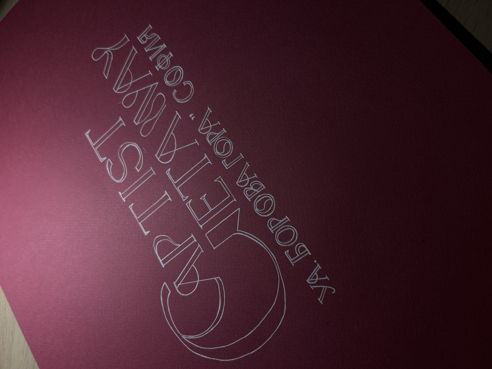The image shows a close-up view of a red or burgundy-colored surface, possibly paper or cardstock, with white lettering and a graphic design. The text, written in a decorative font, appears to be in Bulgarian. It reads "Artist Getaway", followed by "YA Laboratoriya. Sofia". The writing is oriented diagonally across the frame. The background is a solid color, providing contrast to the white lettering. The overall scene looks like a part of an invitation or flyer from an art workshop located in Sofia, Bulgaria.
